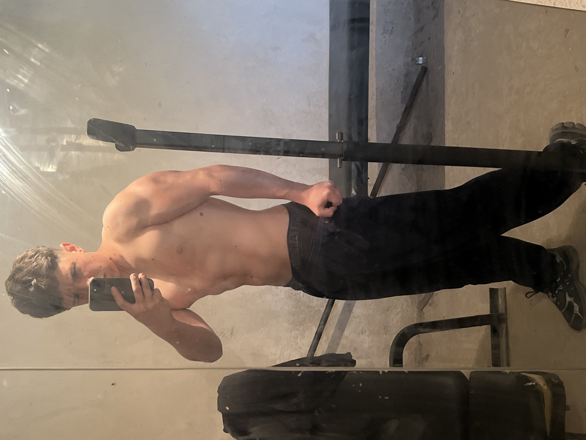 A shirtless man with short brown hair stands facing left, holding a smartphone and taking a selfie in front of a mirror. He is wearing dark pants and athletic shoes. The background includes weightlifting equipment, a concrete floor, and a wall. The location is not identifiable as Zlatograd, Bulgaria, without further information.