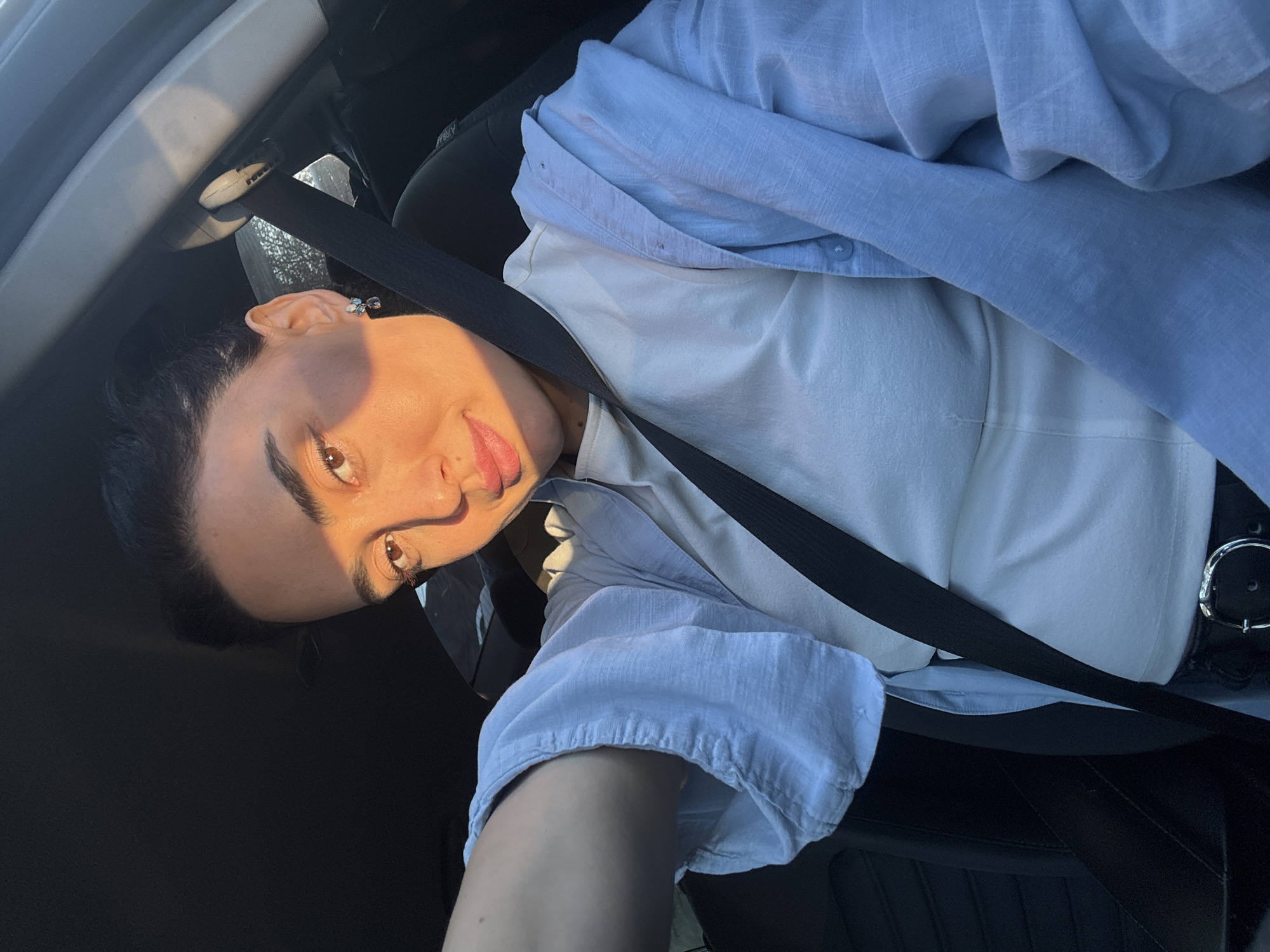 A selfie portrait features a young woman seated inside a car, looking directly at the camera with a playful expression, her tongue slightly visible. Her head is significantly tilted, giving the image a dynamic, rotated orientation. She has dark hair, prominent eyebrows, and brown eyes, with a small catchlight visible in one. Strong, warm sunlight illuminates the right side of her face, casting a distinct shadow on the left and creating highlights on her skin.
She is wearing a light blue, textured button-up shirt unbuttoned over a white or very light-colored top. A black car seatbelt crosses diagonally across her chest and shoulder. Around her waist, a dark belt with a shiny silver buckle is visible. Small, light-colored earrings, possibly shaped like butterflies or bows, adorn her ears. The background consists of the dark, blurred interior of the car, including parts of the seat, headrest, and what appears to be a door panel or roof lining. The dramatic lighting suggests either early morning or late afternoon. The setting is a car, and the provided context places this scene in Tbilisi, Georgia. No readable text is visible in the image.