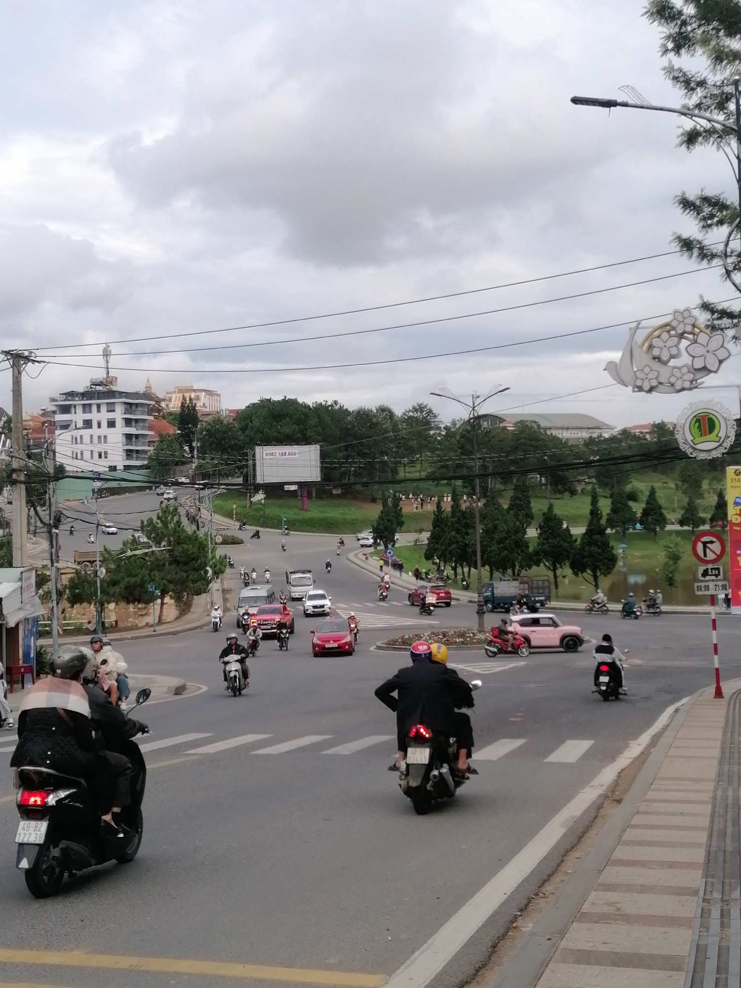 This image captures a busy street intersection or roundabout in Da Lat, Vietnam, under an overcast sky. The scene is dominated by a mix of motorbikes and cars navigating the roads.
In the foreground, several motorbikes are visible, with two prominently featured from behind. The leftmost motorbike has a license plate "49-B2 122.38" and the brand "Lead" on its rear. Another motorbike in the center features the license plate "49-G1 07.844". People on these motorbikes are wearing helmets and casual clothing.
The wider intersection reveals numerous other motorbikes and cars, including a red sedan, a white van, a black pickup truck, and a distinctive pink vintage-style car. Traffic flows around a central median with some greenery. Buildings of varying architectural styles, including a multi-story white building, are visible in the background, nestled among hills and trees. A body of water, possibly a lake or pond, is discernible further back, surrounded by evergreen trees.
Utility poles and numerous power lines crisscross the upper portion of the frame. Streetlights, some with decorative elements resembling leaves or abstract shapes, are also present.
Notable details include a large billboard in the mid-ground displaying the phone number "0982.488.856". A red-and-white road sign on the right indicates "No Right Turn" with the restriction "06:00 - 21:00". Another decorative street sign features a white dove and floral elements, and a partially visible banner appears to have text like "31/4..." and "Ngày...".
The overall impression is that of a typical urban daytime scene in Vietnam, characterized by heavy motorbike traffic and a cloudy day.