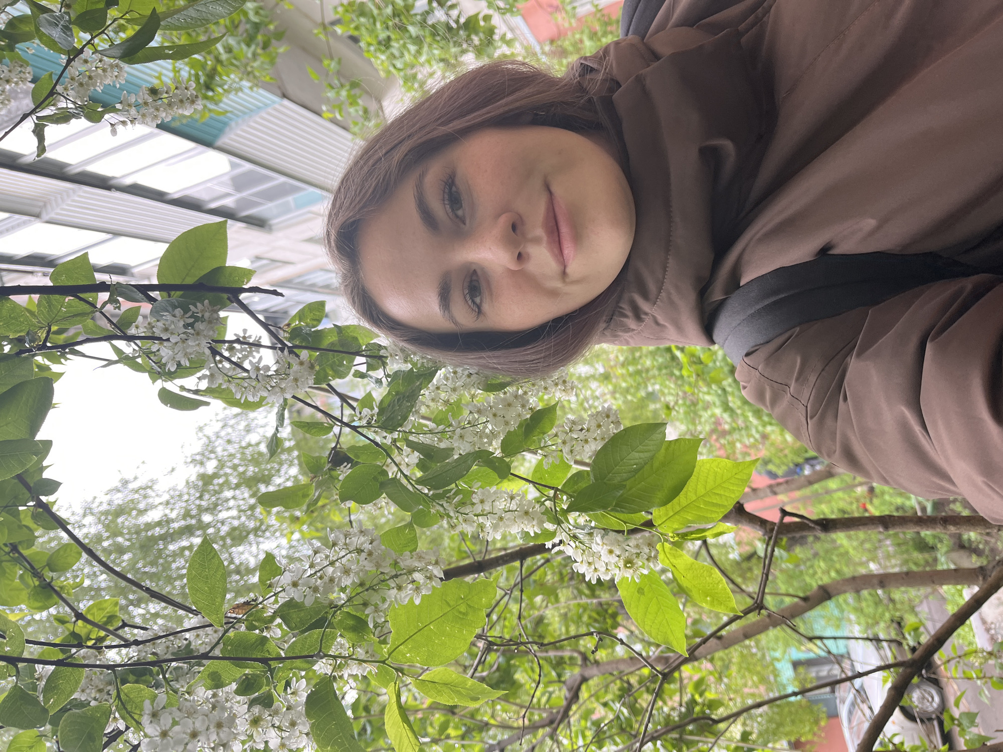 The image is a selfie of a woman with fair skin and brown hair, wearing a brown jacket and backpack. She is standing beneath a tree branch with white flowers and green leaves. The background includes a building with a window facade and patches of sky. The location is identifiable as Chaikovsky, Russia, based on the building architecture visible in the background. The woman is looking directly at the camera with a slight smile.