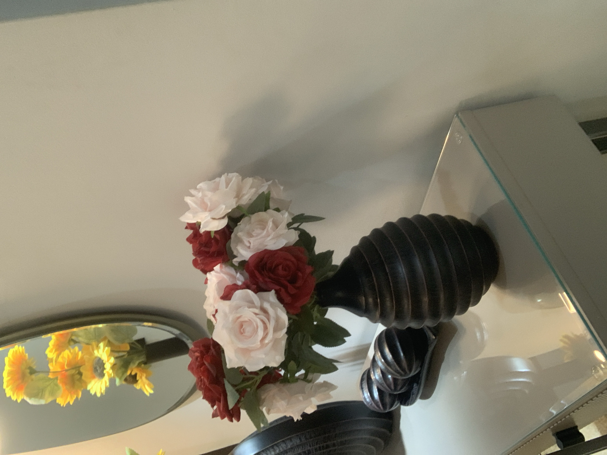 The image shows a floral arrangement of red and white roses in a black vase, placed on a reflective surface, likely a console table with a glass top. To the left of the vase, there are three dark, spherical decorative objects arranged on a tray. In the background, a mirror reflects a bouquet of yellow sunflowers. The overall setting appears to be an interior, possibly a home or a hotel lobby. The wall behind the arrangement is painted a neutral, light color and shows some shadows cast by the objects. The scene is well-lit, highlighting the textures of the flowers and the decorative items. There is no indication of the location being in Abuja, Nigeria, as this is a generic interior scene.