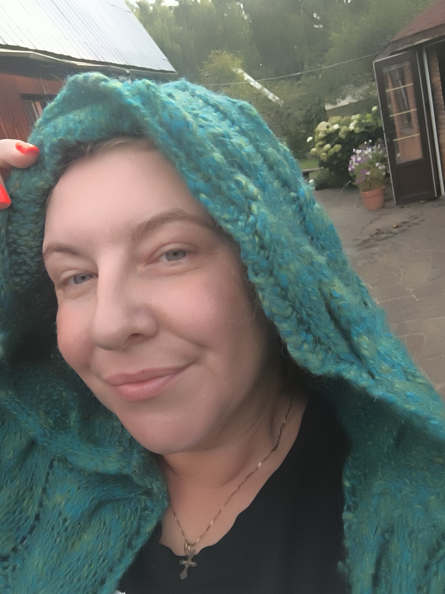 An adult Caucasian female is shown from the chest up, looking directly at the camera and smiling. She has light skin, blue eyes, and light brown hair. She is wearing a thick, knitted teal hooded scarf draped over her head and shoulders. A thin gold chain with a cross pendant is visible around her neck. The background appears to be an outdoor setting, possibly a backyard or rural area. A wooden building with a corrugated metal roof is on the left, and a smaller wooden structure with a glass door is on the right. There is greenery and a potted plant in the background. The lighting suggests it may be daytime, possibly late afternoon or early evening. The image does not provide specific location details within Востец Снт, Russia, beyond the general impression of a residential or semi-rural environment.
