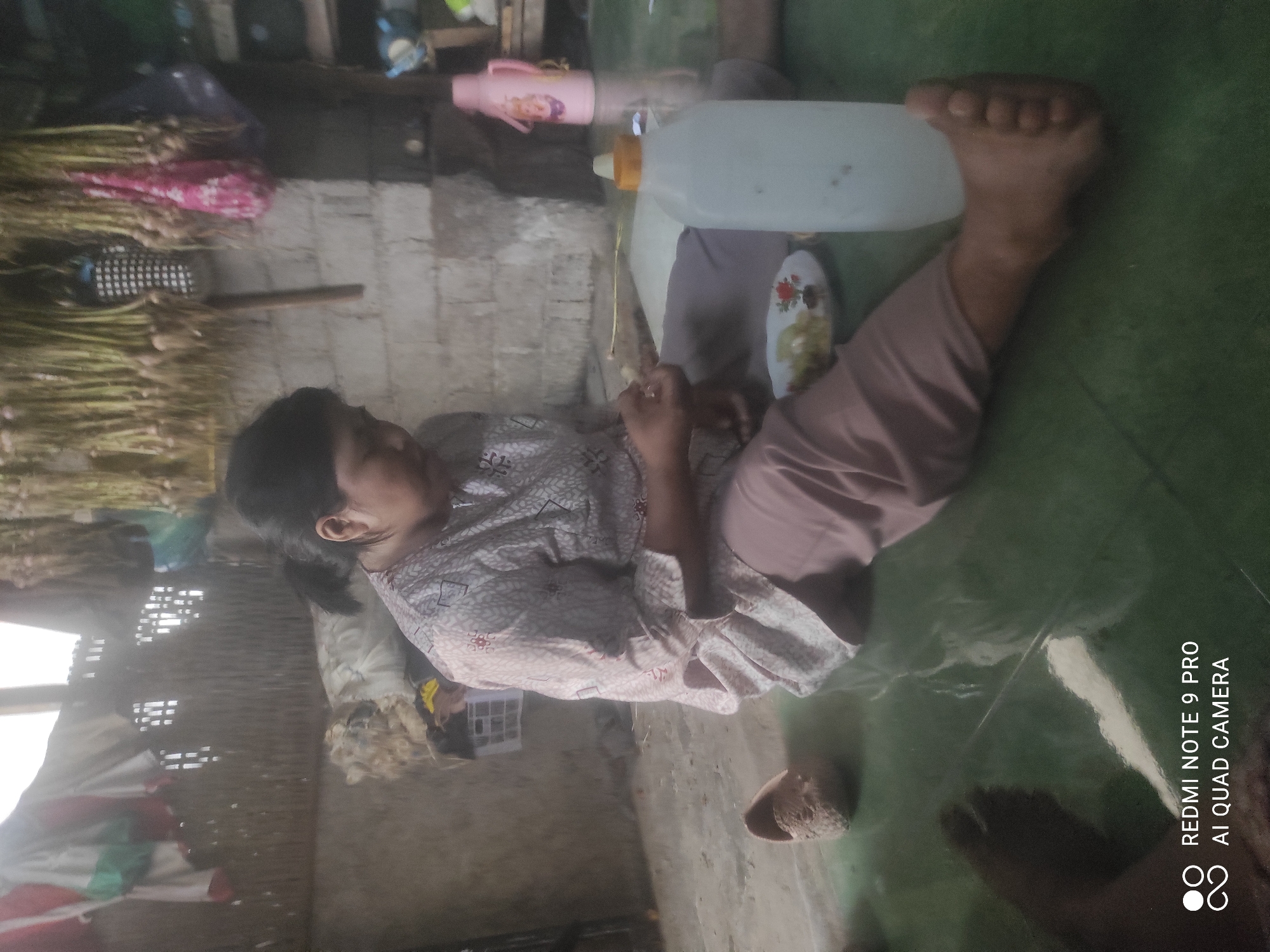 A woman sits on the floor in what appears to be a humble dwelling in Sakra, Indonesia. She is dressed in a patterned shirt and loose pants and is holding a small plate of food. To her right is a large, translucent plastic bottle with a yellow cap, and further to the right, a pink thermos. The background shows a brick wall, a woven matting on the wall, and bunches of garlic hanging from the ceiling. Clothes are hung on a line in the upper left corner. The floor is a green, reflective surface. A single slipper is visible on the floor to the left of the woman.