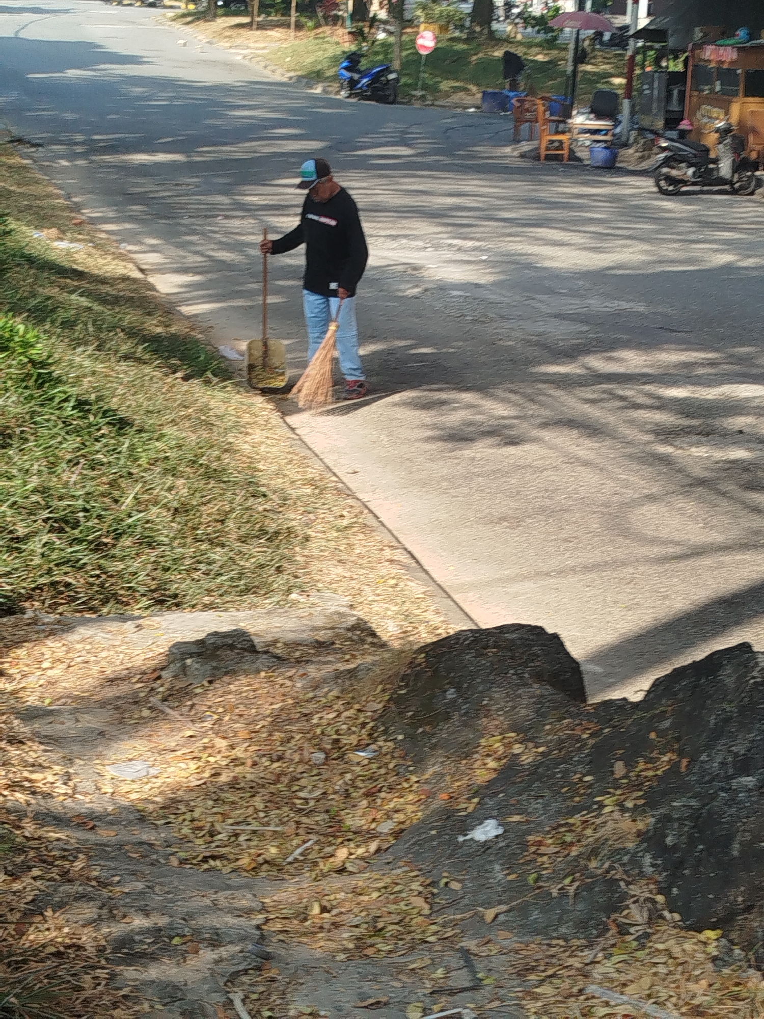 In Balikpapan, Indonesia, a man wearing a long-sleeved black shirt, blue jeans, and a baseball cap is sweeping the side of a road with a broom. A shovel is propped against a pile of debris. The road is paved and lined with grass. Shadows from trees are cast across the pavement. In the background, a food stall, some chairs, and motorcycles are visible. The ground is covered with fallen leaves.