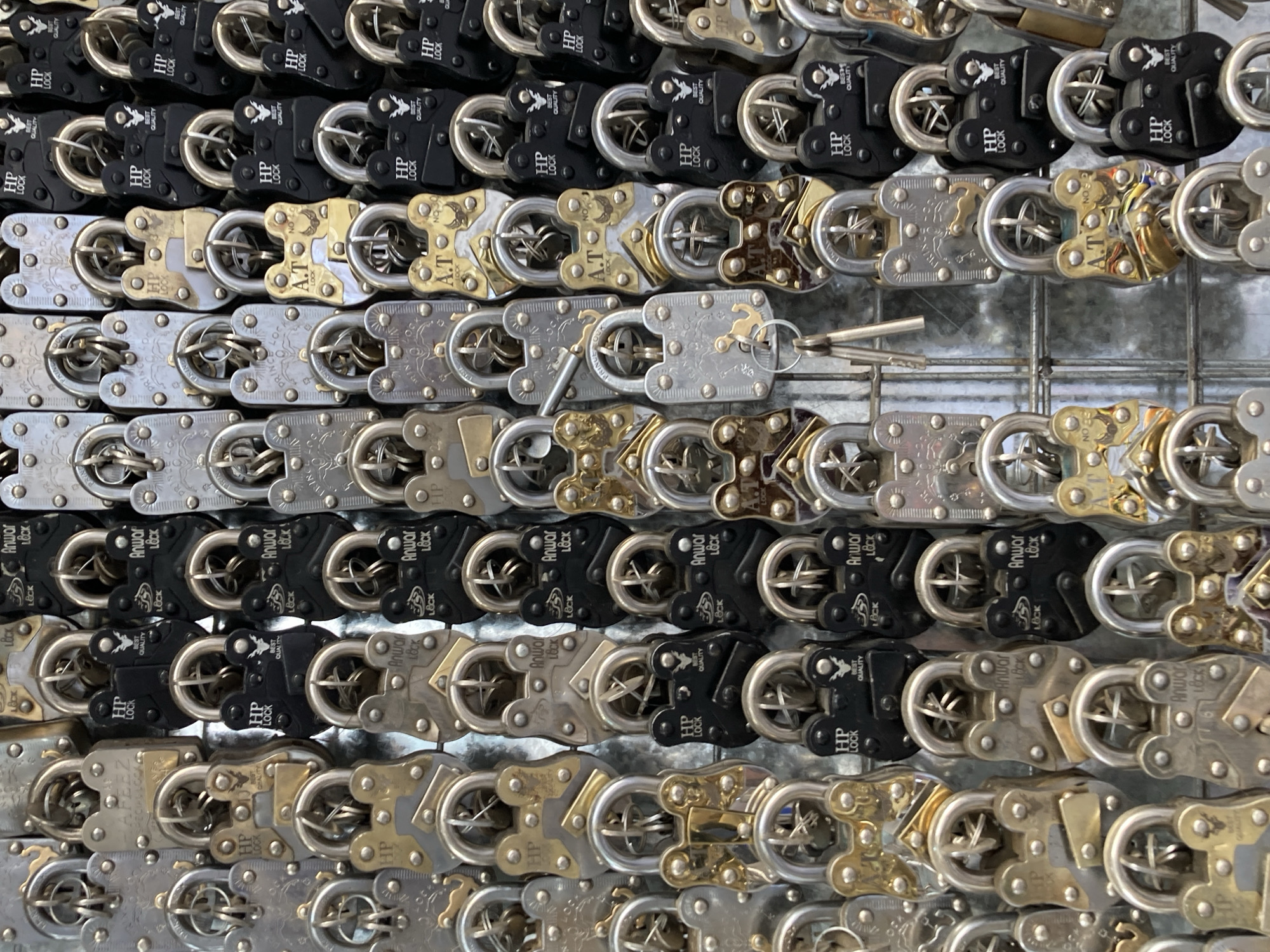 The image shows a close-up of a grid-like arrangement of padlocks, primarily made of silver, gold, and black metals. The padlocks are densely packed together, obscuring what they are attached to. Key brands and names are visible, including "HP LOCK", "Anwar Lock", "A.T Lock," and "AFEEZ PECIALLOCA". Some of the locks have keys attached. The overall impression is that of a display or collection of padlocks. It is speculated that this collection or display is in Quetta City Tehsil, Pakistan based on brands commonly sold there.