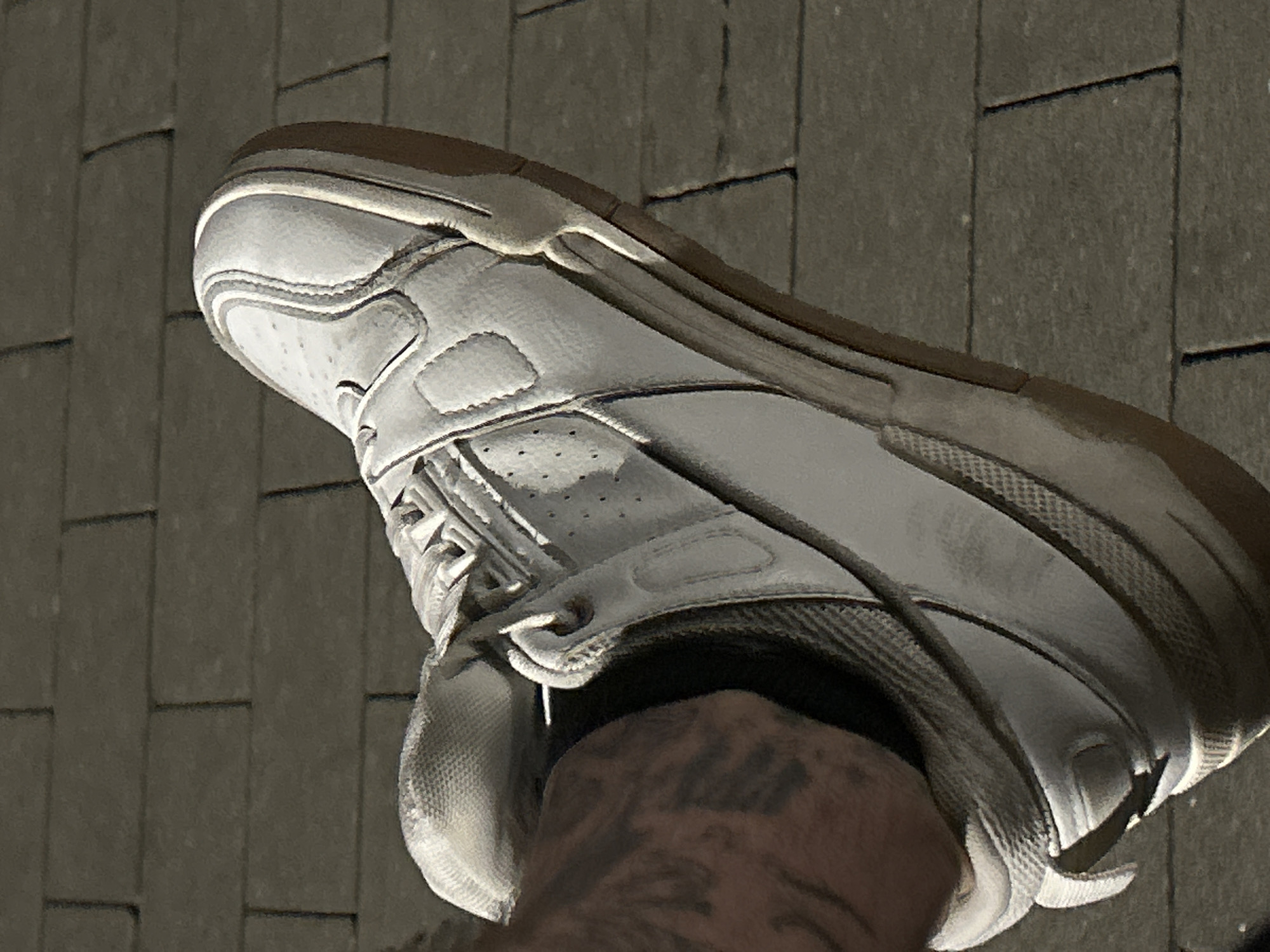 A close-up, low-angle shot captures a white leather sneaker with a gum sole on a person's foot. The shoe is worn, showing scuffs and dirt, particularly on the sole and toe box. The foot is clad in a dark sock, and a tattoo with bold, dark lines is visible on the ankle. The background consists of gray brick paving stones, casting distinct shadows, indicating the presence of sunlight. This image is not set in Siedlce, Poland; it is a generic urban outdoor setting.
