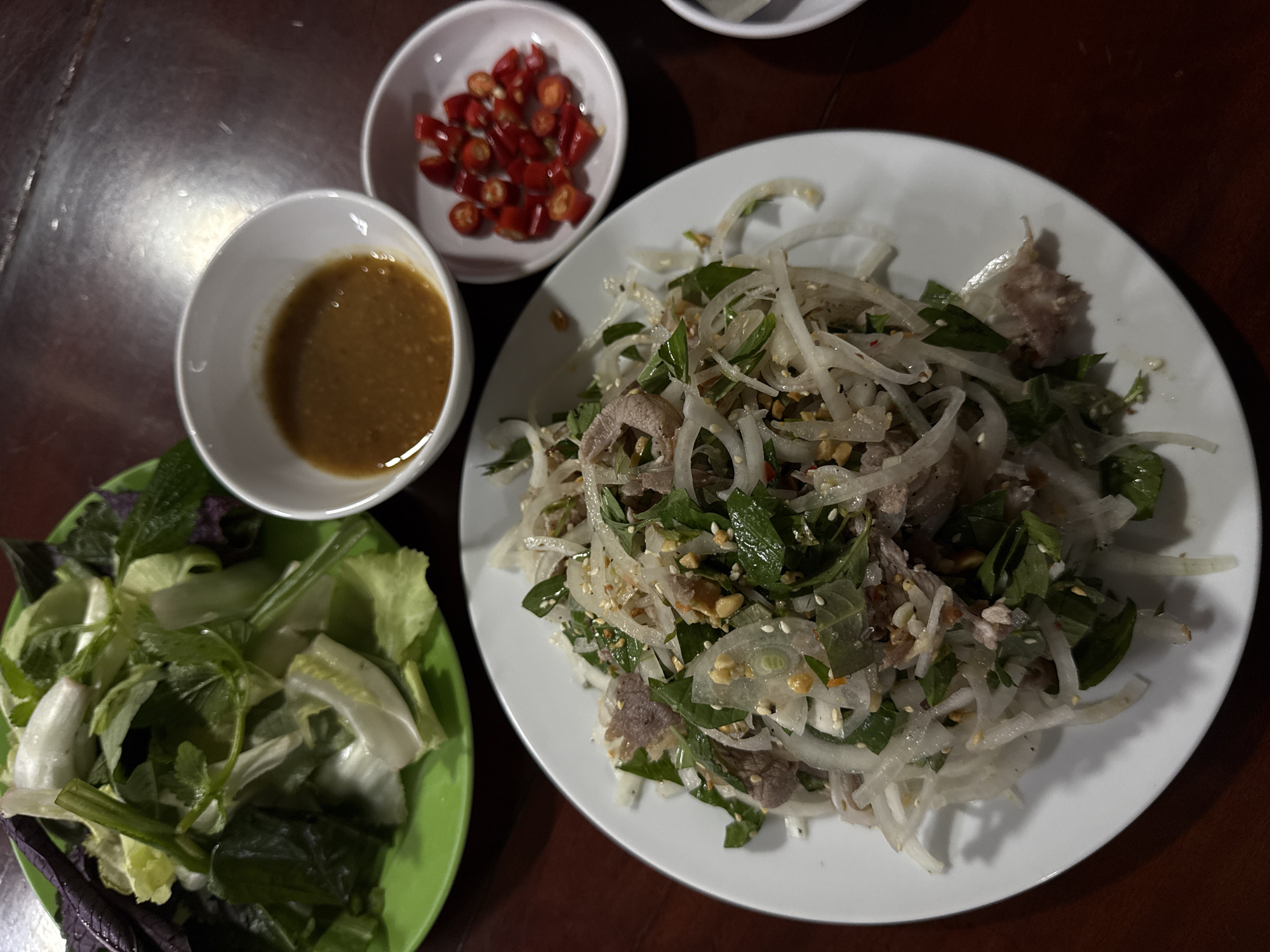 A high-angle shot shows a meal on a wooden table. In the foreground, a white plate holds a salad with thinly sliced meat, onions, herbs, and sesame seeds. To the right of the salad, a small white bowl contains chopped red chilies, and further to the right, partially visible, is another small white bowl, possibly holding a dipping sauce. In the background, a green plate is filled with a variety of fresh green vegetables and herbs. The scene is set in a restaurant in Da Lat, Vietnam.