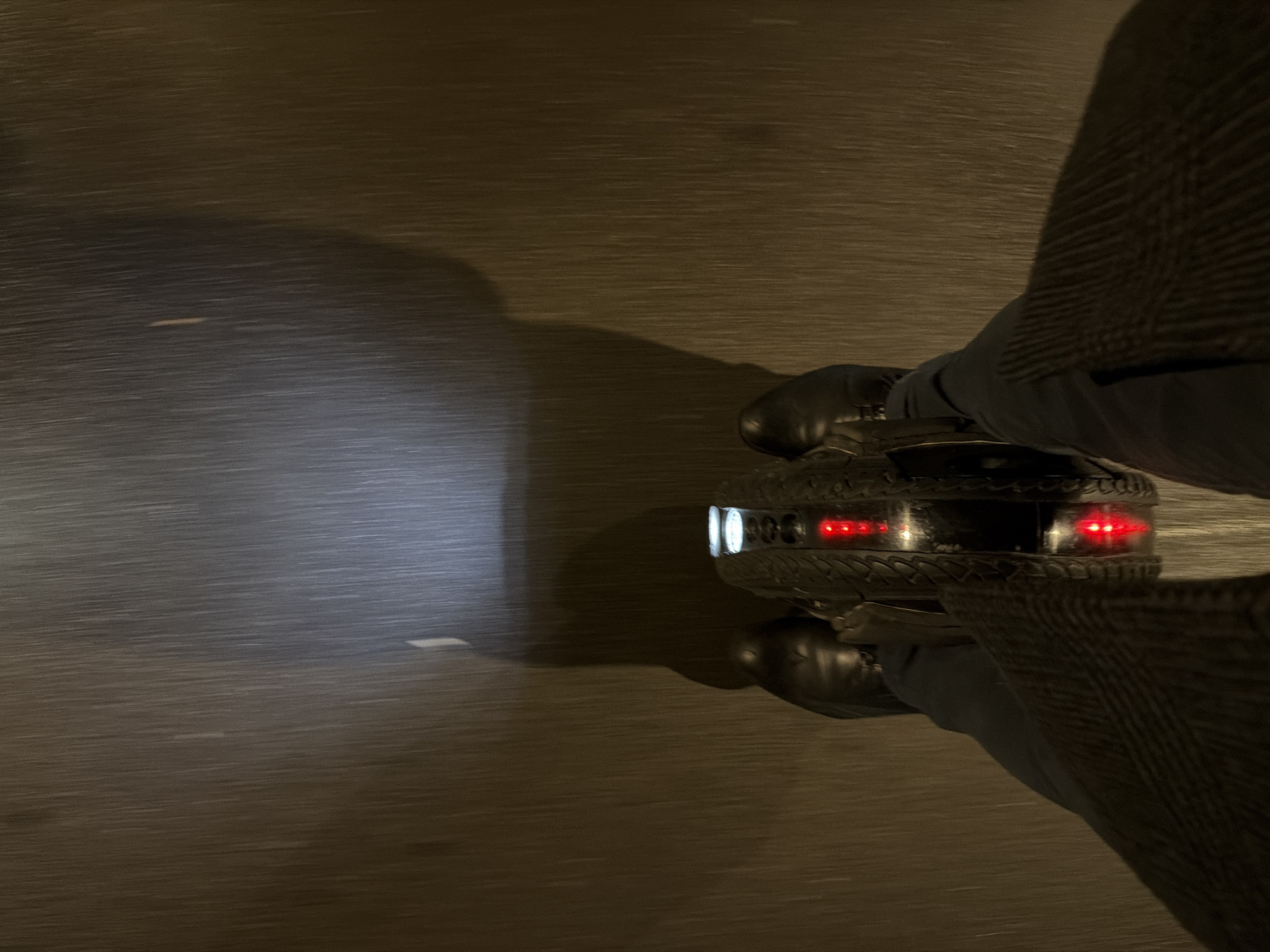 A person is riding an electric unicycle on a paved surface at night. The unicycle has a white headlight and red taillights, both illuminated. The person is wearing shoes and dark pants. The background is blurred, suggesting motion. The location cannot be determined from the image.