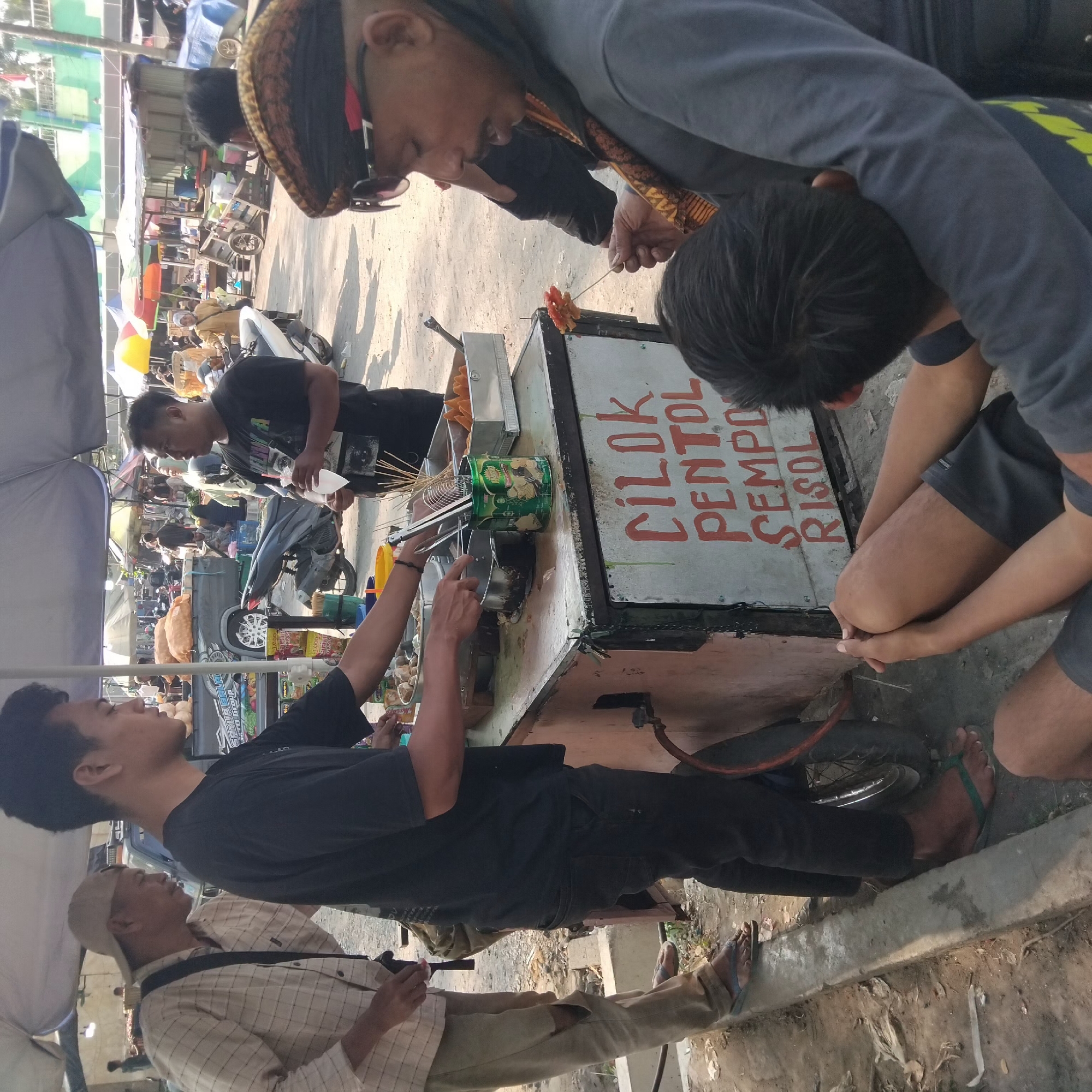 The image depicts a street food vendor in Banjarbaru, Indonesia. A man, likely the vendor, is preparing food at a cart. He is wearing a black t-shirt and dark pants. In front of him, on the cart, are various food items, including what appear to be meatballs and fried snacks on skewers. The cart has text written on its side that reads "CILOK PENTOL SEMPOL RISOL," identifying the food being sold.
To the left of the vendor, an older man wearing a light-colored plaid shirt, beige pants with a ripped knee, and a tan baseball cap stands observing. He has a bag slung across his body.
In the foreground and to the right, a man with a shaved head, wearing sunglasses and a traditional patterned head covering (iket), is holding a skewer with food. He is looking down at the skewer. Another individual, whose face is not visible, is seated in the lower right corner, with their arms around the seated person.
The environment suggests a bustling outdoor market or street setting. In the background, other vendors, stalls, and people are visible, along with parked motorcycles and vehicles, indicating a lively commercial area. The ground is unpaved, and a concrete curb runs along the left side of the frame. The overall lighting indicates it is daytime.