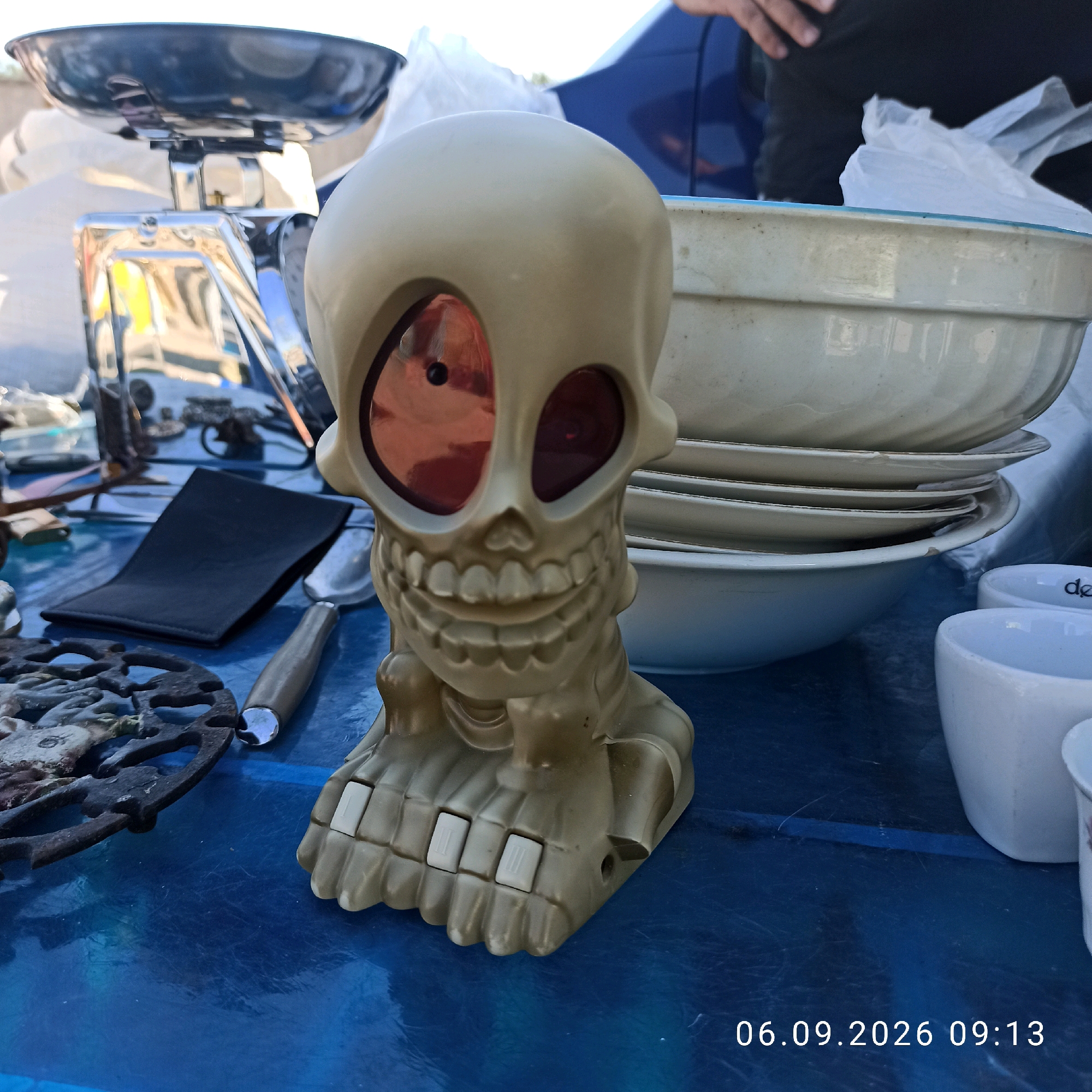 The image shows a close-up of a table with various items, possibly at an outdoor market. In the foreground, a plastic skull decoration with red eye sockets and a grinning mouth is prominent. Its base features several white squares. To the left of the skull is a black, folded object, possibly a wallet or case. A metal object with a decorative pattern, resembling a trivet, is also visible. Behind the skull, a stack of white bowls and plates are arranged. A chrome scale with a large metal basin sits further back on the left. A hand, partially visible at the top right, suggests a person is present. The background includes a blue car and some white plastic bags. The overall surface is a blue tablecloth or tarp. The date stamp on the image reads "06.09.2026 09:13." The scene does not provide any specific visual cues to pinpoint the location to Sannicandro di Bari, Italy.
