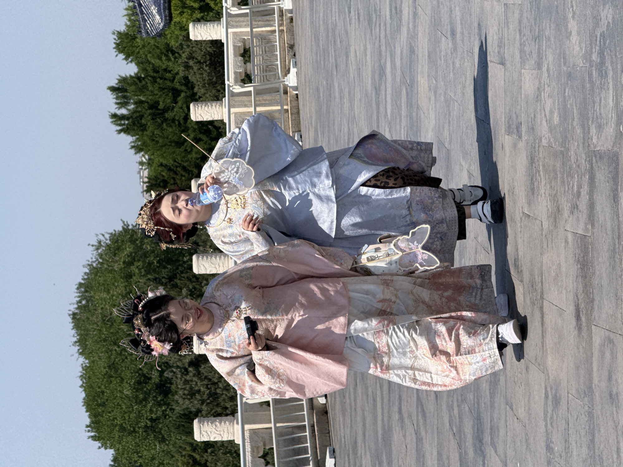 Two people wearing what appears to be traditional Chinese clothing are standing on a stone walkway. The person on the right is holding a water bottle to their lips. The person on the left is holding a small black camera. Behind them is a manicured green space and a series of white balustrades. The ground is paved with rectangular stones. Based on the architecture, this is likely in the city of Dongcheng District, China.