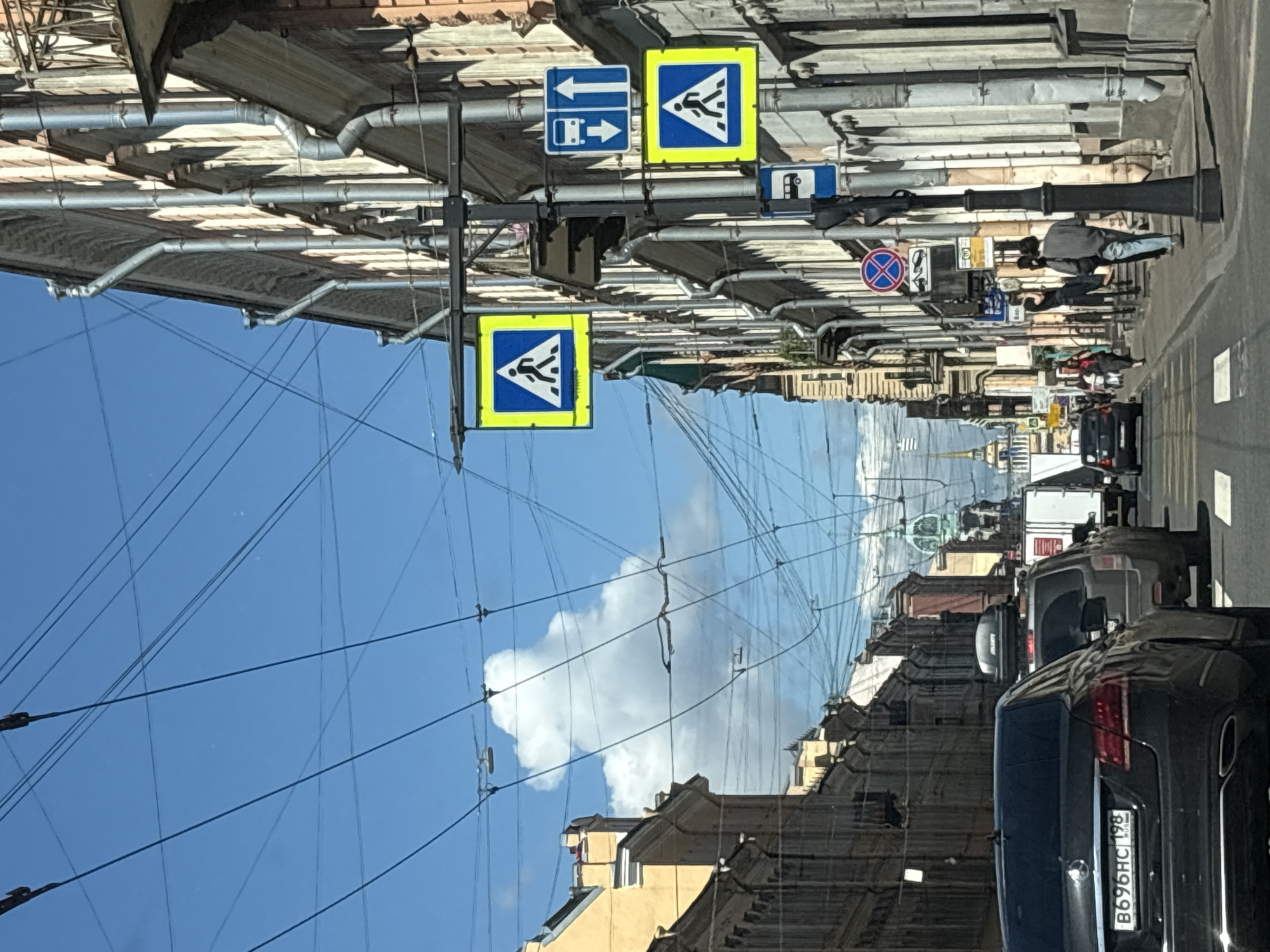 The image shows a street scene in Saint Petersburg, Russia, likely on a sunny day. The perspective is from inside a vehicle, looking forward. The street is lined with buildings exhibiting classic Saint Petersburg architecture, characterized by their ornate facades and multiple stories. Several vehicles are visible on the road, including a dark Mercedes-Benz sedan in the foreground with a Russian license plate. Overhead, a complex network of electrical wires crisscrosses the sky, characteristic of older city infrastructure.
Traffic signs are prominent. Two yellow pedestrian crossing signs, featuring a white silhouette of a person walking, are mounted on a pole. Another blue sign displays an arrow pointing upwards and a pictogram of a bus, indicating bus lane or route. A circular red and blue sign with a diagonal line through it, likely a prohibition sign, is also present.
People are seen walking on the sidewalk. Some are interacting, with two individuals standing near a lamppost, one in a grey sweatshirt and jeans, the other in dark clothing. Further down the street, more pedestrians are visible, and a white truck is parked. In the distance, a spire or tall building with a greenish dome is visible, a common feature of Saint Petersburg's skyline. The sky is a bright blue with scattered white clouds. The overall impression is of a bustling urban environment with a blend of historical architecture and modern life.