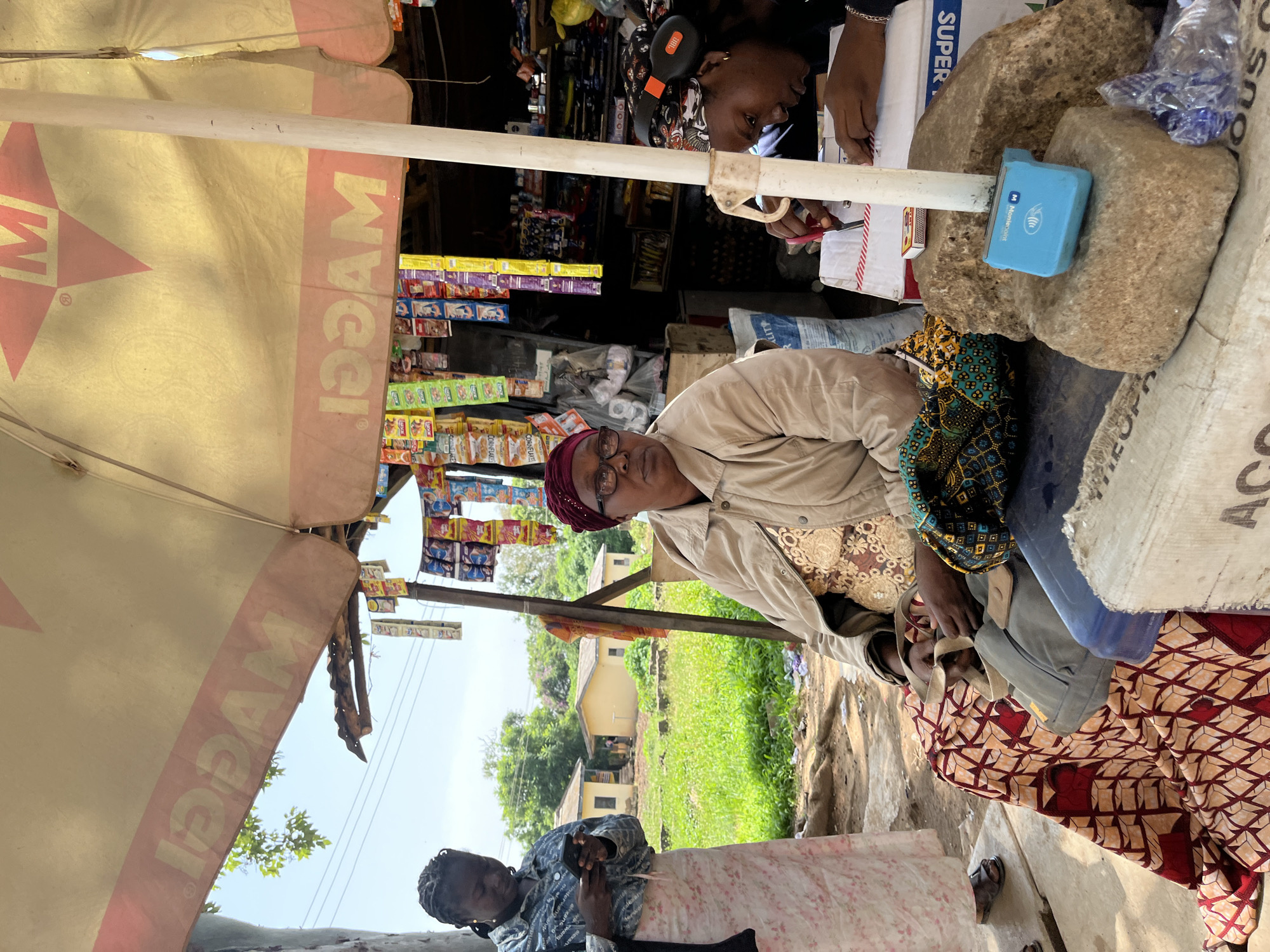 This image depicts an outdoor market scene in Rafin Par, Nigeria. In the foreground, a woman wearing a patterned headwrap and glasses sits under a large yellow and pink umbrella that has "MAGGI" branding. She is wearing a light-colored jacket over a gold and white patterned garment. She is holding a piece of fabric in her hands. To her right, another woman, wearing a headscarf with a patterned print and headphones, is seated and appears to be writing on a piece of paper. In front of them are several large rocks, one of which holds a small blue electronic device. A box with "SUPER" printed on it is also visible. To the left of the seated woman, another woman stands holding a smartphone. She is wearing a denim-like shirt and a long, light-colored patterned skirt. In the background, there are small yellow buildings and lush green vegetation under a bright sky. Stalls with various packaged goods, likely food items, are set up under the umbrella. The overall scene suggests a bustling marketplace environment.