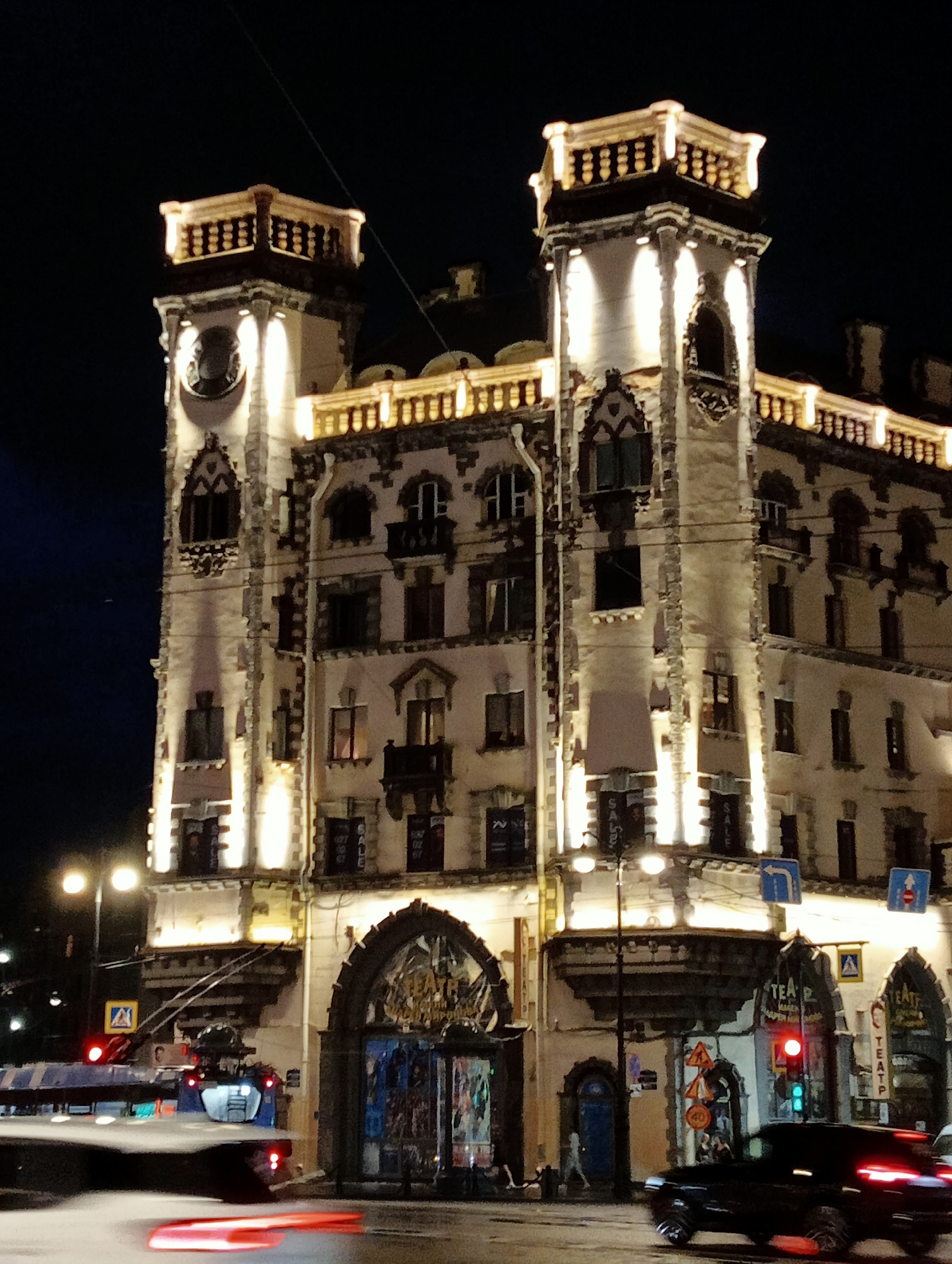 An illuminated building, likely a historical landmark in Saint Petersburg, Russia, stands prominently at night. The architecture features ornate detailing, including two prominent towers with decorative railings at the top, and arched windows. The building is lit by numerous spotlights, casting a warm glow against the dark night sky. Several windows are visible, some appearing lit from within.
In the foreground, a street scene unfolds. A blue trolleybus is partially visible on the left, its red taillights creating a streak of light due to motion blur. A black car, also with illuminated taillights, is driving away from the camera on the right. Traffic lights are visible, one showing a red light. Street signs and traffic signs are scattered along the street. The ground appears to be wet, reflecting the lights. The overall impression is a vibrant urban nightscape of a significant city.