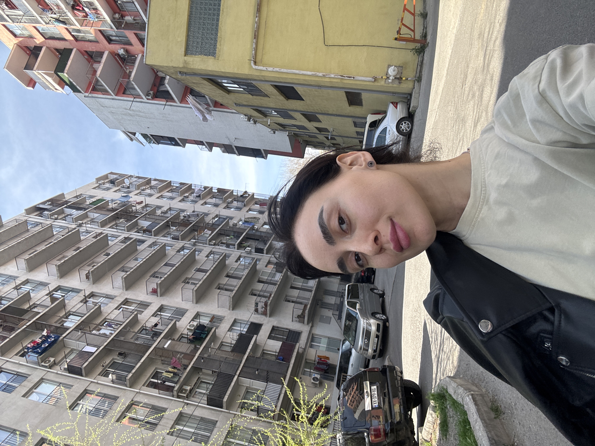 A young adult poses for a selfie, standing outdoors with her head tilted to the right. She is wearing a light-colored t-shirt and a black leather jacket. Behind her are tall, Soviet-era residential buildings, common in Tbilisi, Georgia. Clothes are hanging on some balconies. Cars are parked on the street in the background. A section of yellow building is also visible. The lighting suggests it's daytime.