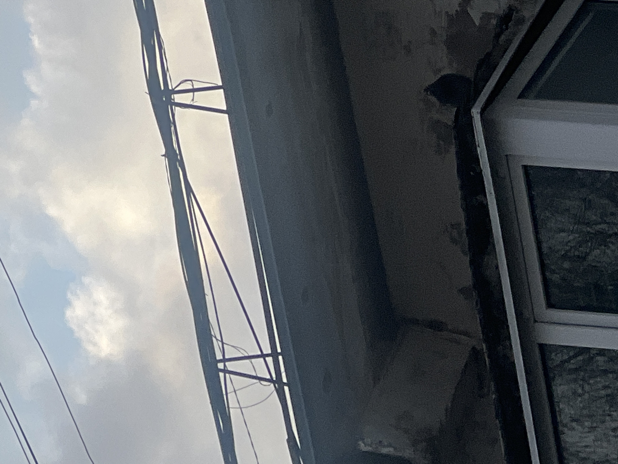 The image shows an upward angle view of a building facade, possibly located in Chaikovsky, Russia, as requested, but without visual confirmation of the location.
The building features a window with a white frame and panes reflecting an unclear exterior scene. The facade is aged, with peeling paint and signs of weathering. Utility lines are visible against the sky, which shows a cloudy sky. The lines appear to be connected to the building with support structures.