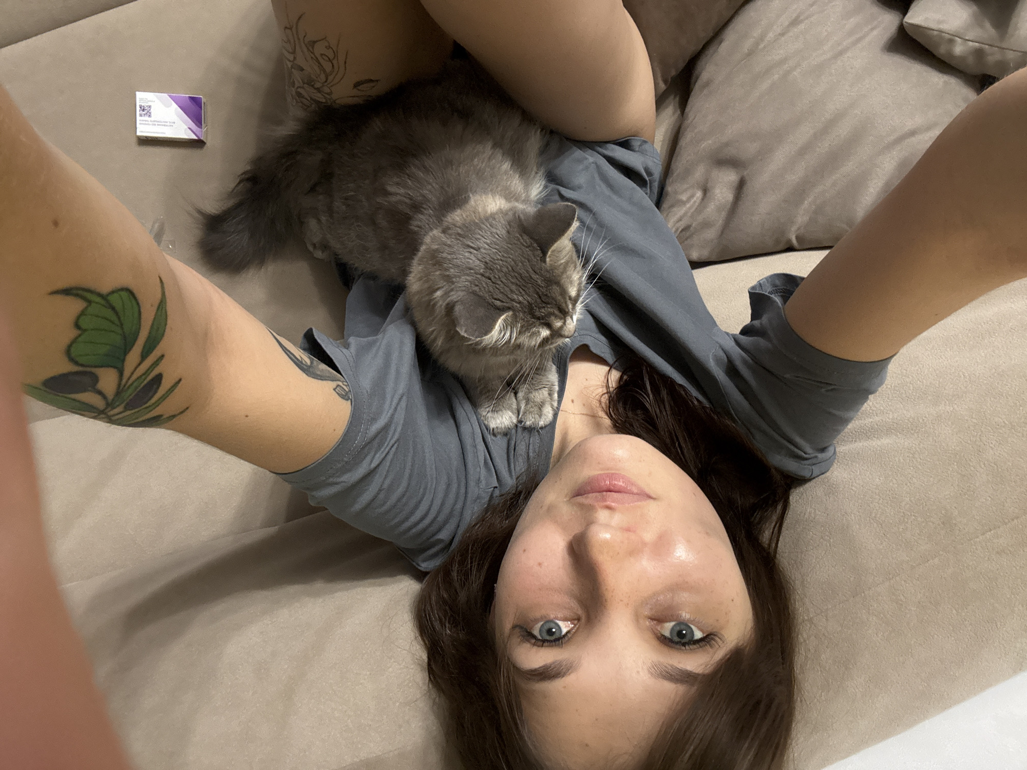 A person is lying on a beige sofa, holding a grey cat. The person is looking at the camera, with their eyes open and a slight smile. They have brown hair and blue eyes, and are wearing a grey t-shirt. The cat is resting on the person's chest, with its head tilted back and its eyes closed. There are tattoos on the person's arm and leg. A small white and purple box is placed on the sofa next to the person. The scene appears to be indoors, likely in a residential setting. There is no information to suggest the location is Novokuznetsk, Russia.