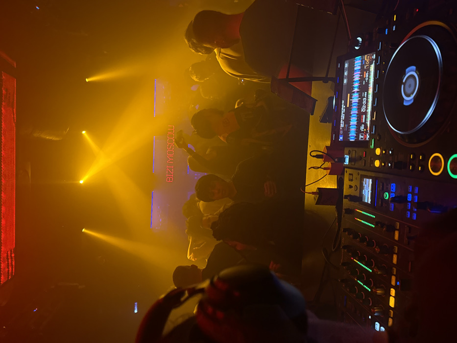 The image captures an indoor nightclub environment at Bizi Moscow in Moscow, Russia. In the foreground, a DJ console is visible, comprising a multi-channel mixer with illuminated green and blue faders and buttons, and at least one digital media player (CDJ) displaying waveform information on its screen, with a rotating platter. A pair of over-ear headphones rests on the far left. On the table behind the DJ equipment, two power adapters (one black, one red) with connected cables are present, along with a tablet stand. A person's forearm and hand are partially visible on the left, operating the mixer.
The midground and background feature a dense crowd of individuals, estimated at two to three dozen, standing and interacting in a dimly lit, hazy atmosphere. Their silhouettes are prominent against the intense orange-yellow light beams originating from overhead spotlights. A prominent red neon sign in the background spells out "BIZI MOSCOW", flanked by horizontal blue light strips. The overall scene indicates an active social gathering or performance event within the described venue.
