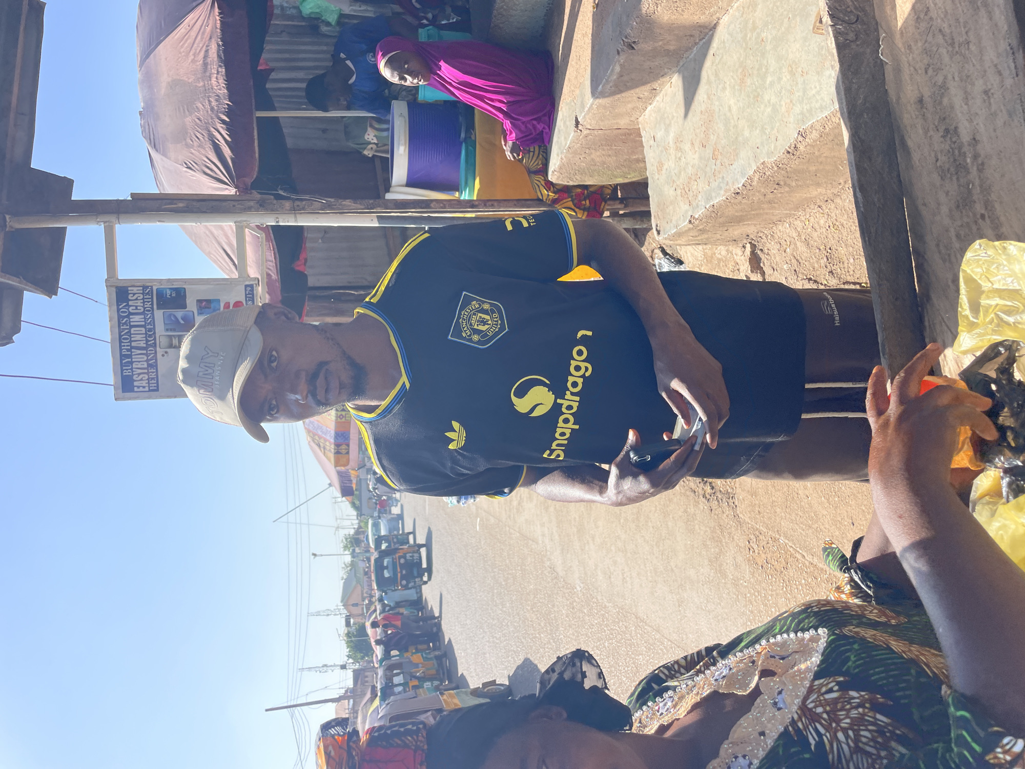 In Gura Top, Nigeria, a street market scene unfolds under a bright, clear sky. In the foreground, a man wearing a black Manchester United jersey with yellow trim and a beige baseball cap stands looking towards the camera. He holds a mobile phone and a small white object in his hands. To his left, a woman in a green and brown patterned dress with elaborate embellishments around the neckline is partially visible, her hands in front of her, seemingly engaged in handling items on a wooden surface. Behind them, a woman in a vibrant pink hijab is seated on concrete steps, looking towards the right. Further in the background, a bustling street is lined with various vehicles, including auto-rickshaws and cars, and stalls with umbrellas providing shade. A sign board advertising "BUY PHONES ON EASYBUY AND IN CASH HERE AND ACCESSORIES" is prominently displayed. The overall environment suggests a lively commercial area with a mix of vendors and shoppers.