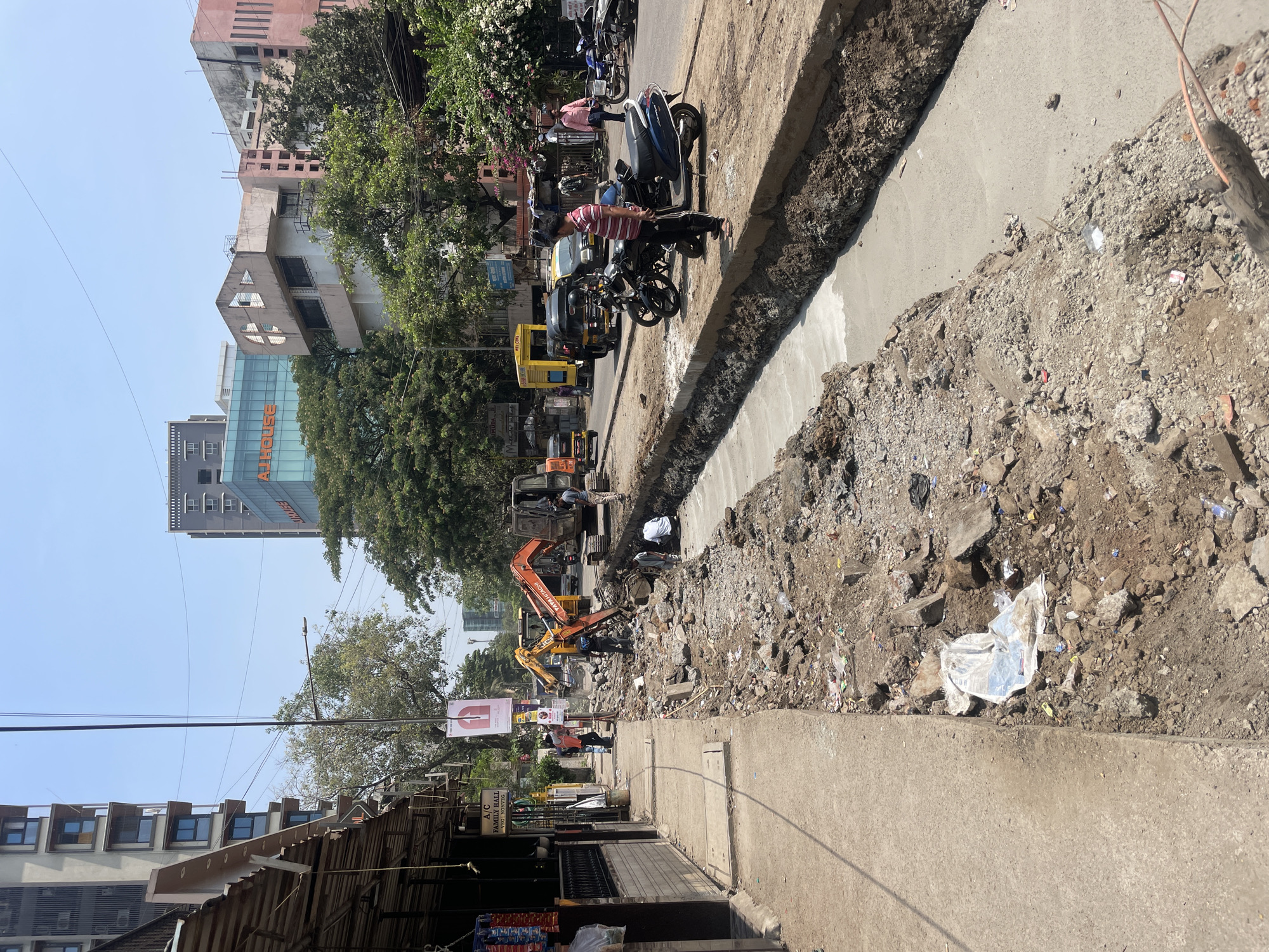 The image depicts a street scene in Mumbai, India, undergoing construction. A deep trench runs along the side of the road, filled with excavated material, debris, and construction equipment. An orange excavator is visible within the trench, seemingly digging or performing related tasks. Further down the road, yellow auto rickshaws and motorcycles are parked. Pedestrians are present on the sidewalk and along the street. Buildings are visible in the background, ranging from residential structures to a high-rise with the word "All House" on it. The weather appears to be sunny and clear.