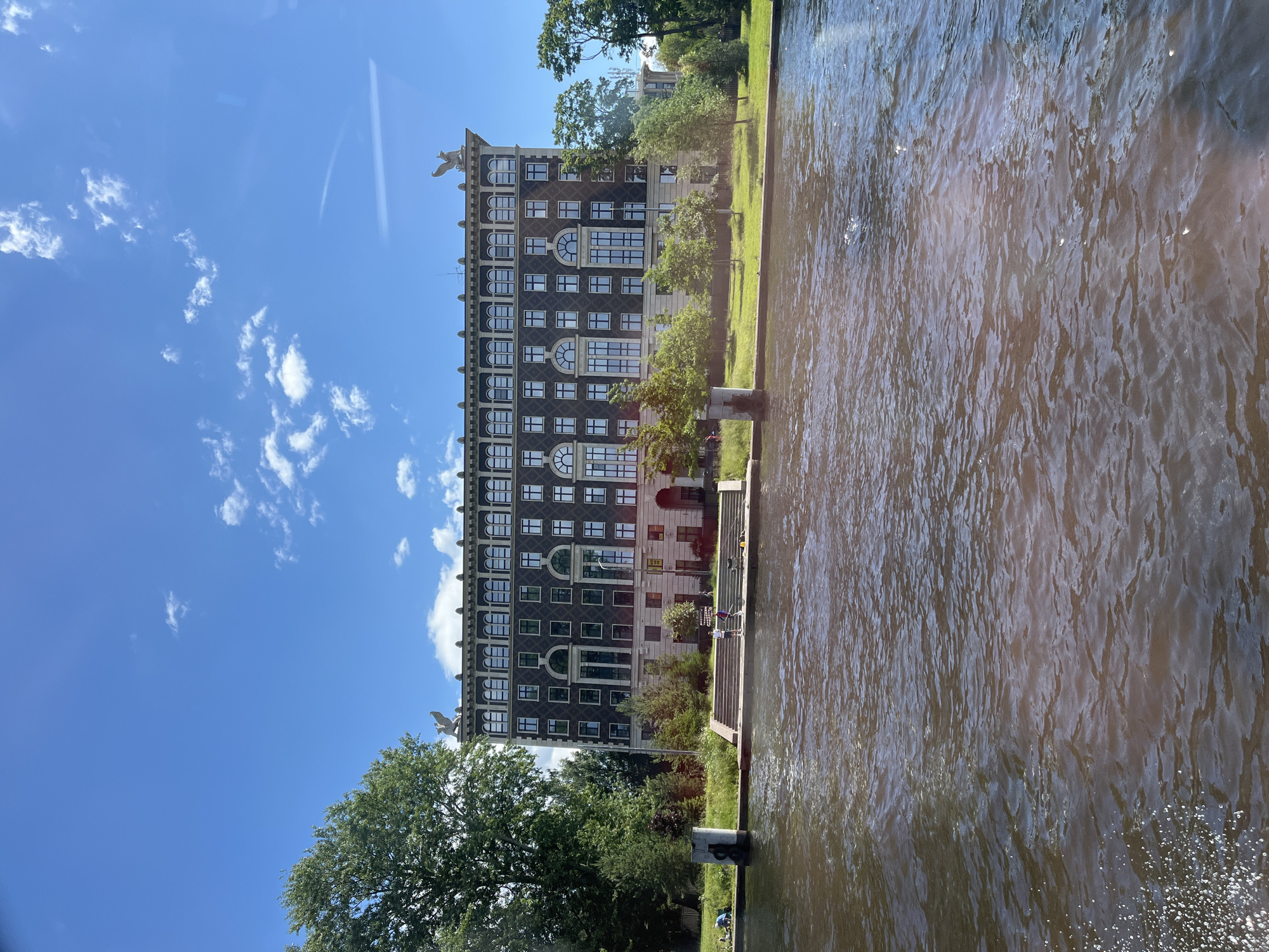 A large, multi-story building with a dark facade and numerous windows stands on the bank of a wide body of water. The building has decorative elements, including what appear to be griffins on the roofline. In front of the building is a grassy area with trees and a set of stairs leading down to the water's edge. Several people are visible on the stairs and on the grassy area, some appearing to be engaged in leisure activities. The water in the foreground is rippling. The sky above is blue with scattered white clouds. The building is likely the Narva Power Plant (Narvskaya GRES), located on the Neva River in Saint Petersburg, Russia, although it is atypical for this building to be seen from this perspective without the presence of the power plant's chimneys.