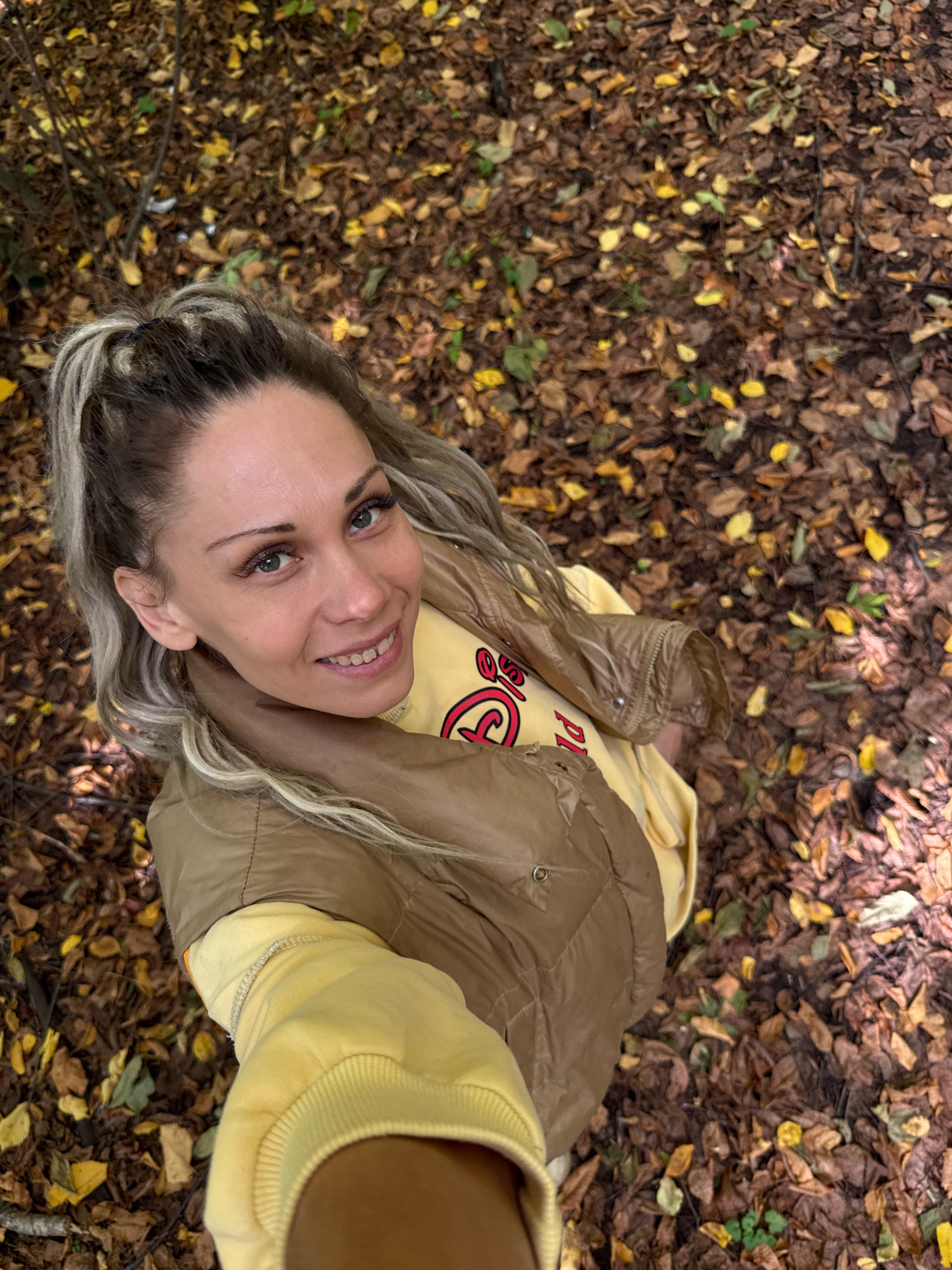 A high-angle, medium shot captures a woman in a park setting. She is wearing a tan puffer vest over a yellow sweatshirt, and her blonde hair is pulled into a high ponytail. Her eyes are looking at the camera, and she has a smile on her face. The ground around her is covered in fallen leaves. The image appears to be a selfie, taken outdoors in what is likely a wooded area with fallen leaves on the ground. The setting is consistent with a park in Rusanovo, Russia during the autumn season.