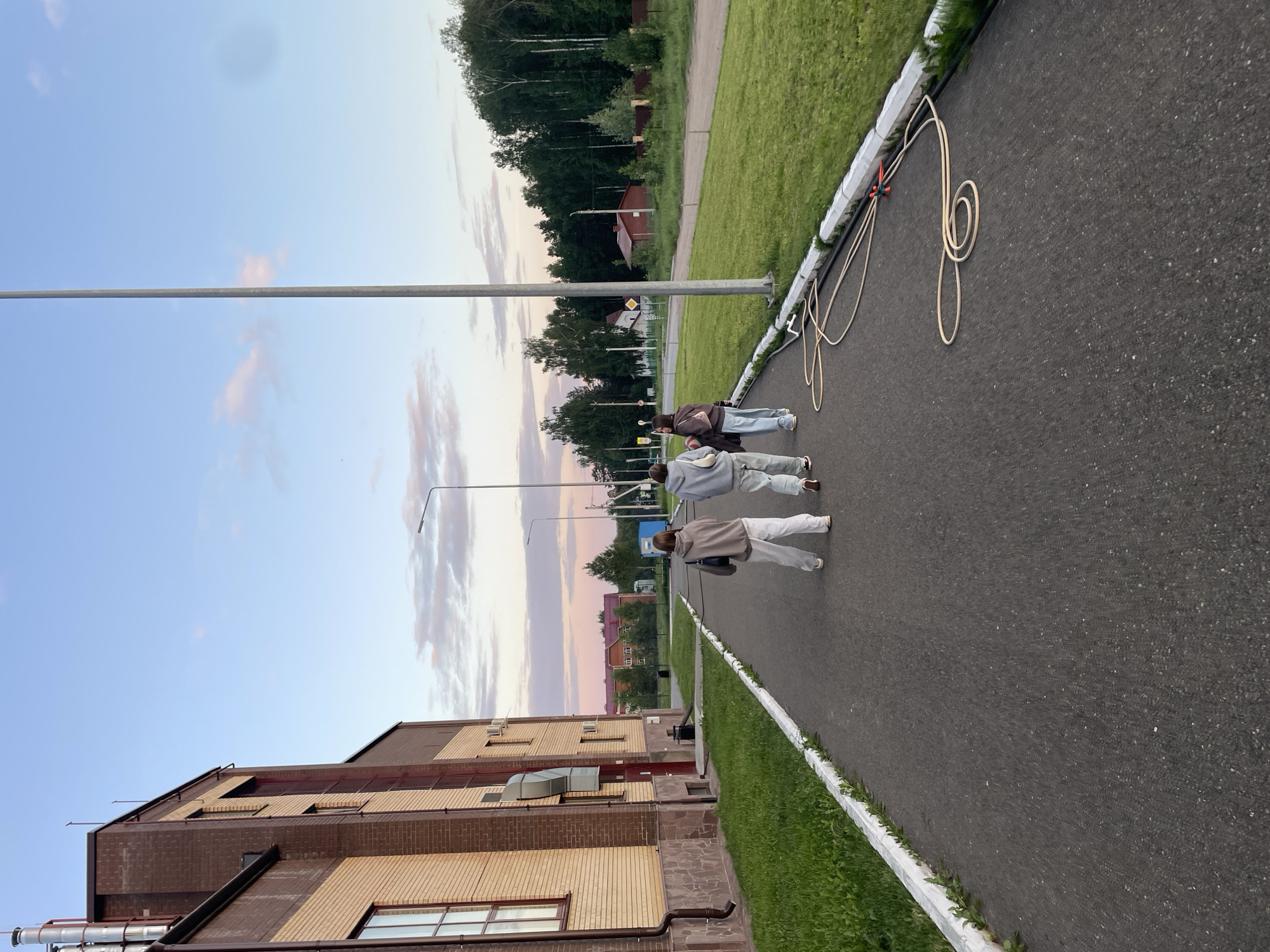 An outdoor scene in Noviy, Russia, captures three individuals walking away from the camera on a dark asphalt path during dusk. The sky above is light blue with scattered pink and white clouds, indicating either sunset or sunrise, with soft, ambient lighting.
On the left, a multi-story brick building stands, featuring a base of dark reddish-brown bricks and a lighter beige brick facade on its upper sections. The building has several dark-framed windows, vertical dark panels, exposed metallic pipes, and external ventilation units. Green grass borders the left edge of the path.
The asphalt path extends into the distance, with a white concrete curb marking its right edge. Alongside this curb, there is a strip of green grass, followed by a wider expanse of neatly mown grass. A tall, slender metal pole, likely a streetlight, rises on the right side of the path, partially cropped at the top. Near the right curb, a beige hose is coiled on the asphalt, connected to some black tubing and an orange plastic splitter/connector.
The three individuals, seen from behind, are walking in a staggered line down the path. The person on the far left wears light-colored pants and a dark grey hoodie, carrying a dark bag on their left shoulder. The middle person wears similar light-colored pants and a lighter grey hoodie. The person on the right wears light blue jeans and a dark top, carrying a brown backpack.
In the mid-ground and background, dense forest with tall trees forms a prominent backdrop on the right. Between sections of trees, several distant buildings are visible, including a small red-roofed structure and other residential-style buildings. Another streetlight is visible further down the path on the right.