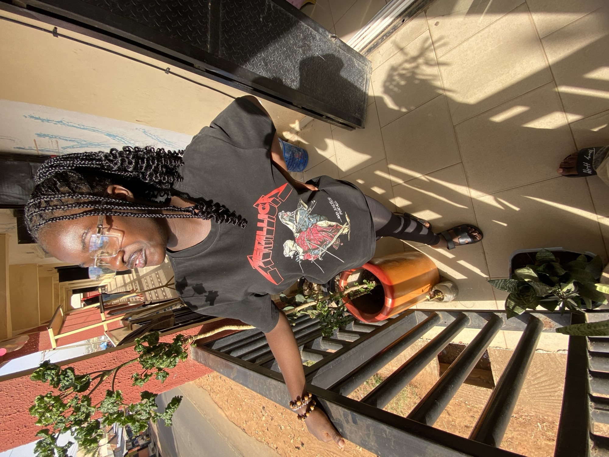 A woman stands on a tiled patio overlooking a street in Bukuru, Nigeria. She wears glasses, braids, a black t-shirt with a Metallica graphic, and black leggings. She leans against a black metal railing with her right hand, adorned with beaded bracelets. Below the railing is a street. On the patio are potted plants, including a snake plant and another in a reddish-brown pot. A pair of sandals lies on the tile near the plants. A dark shadow suggests overhead foliage. A blue object is visible on the patio near a metal door or gate in the background.