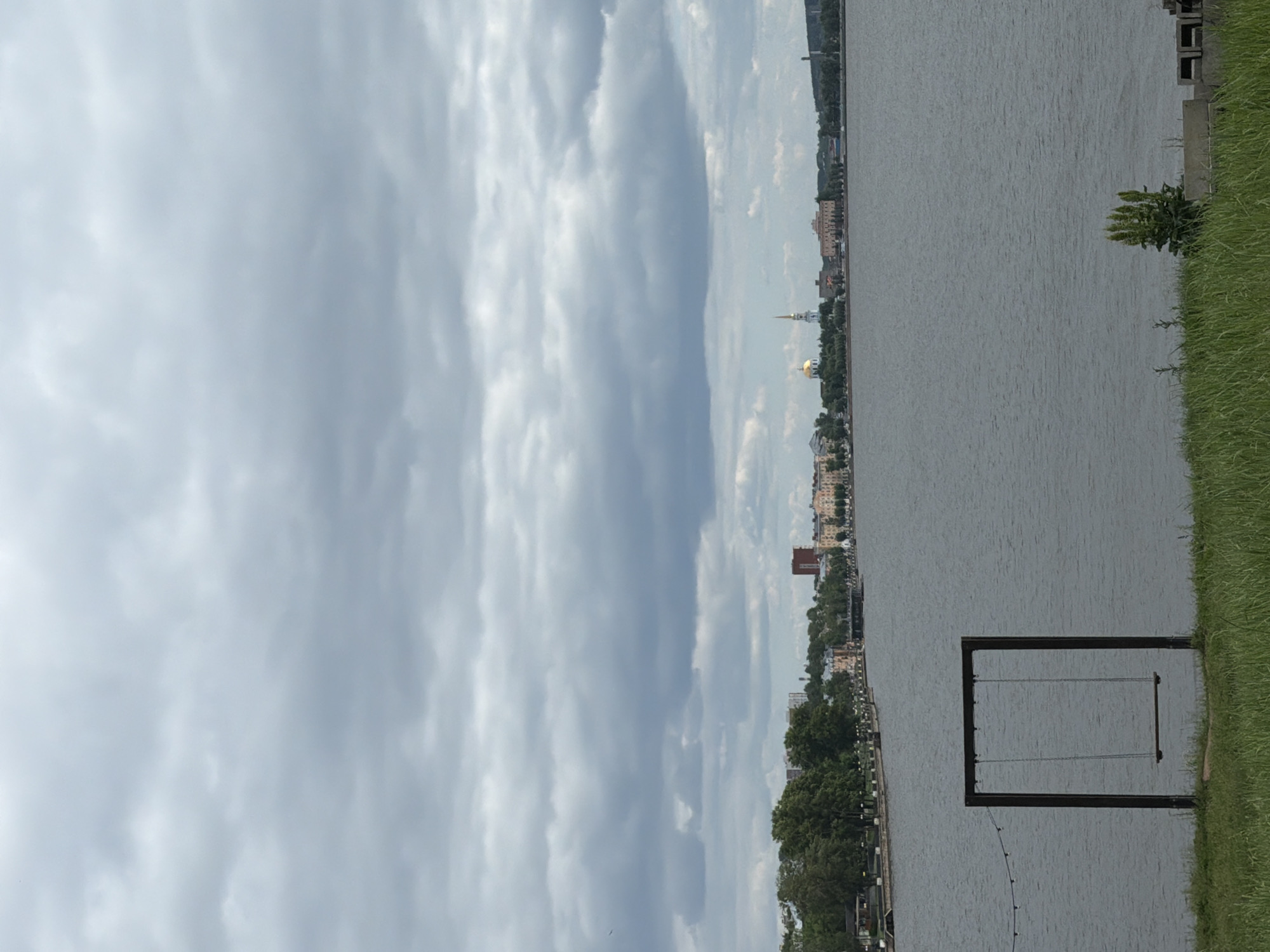 The image depicts a serene waterfront scene in Votkinsk, Russia, under a cloudy sky. In the foreground, lush green grass leads to a solitary, dark-framed swing set overlooking a calm body of water. The middle ground features a distant cityscape with a variety of buildings, including a prominent church with a golden dome and a spire, suggesting a historical or religious significance. Trees line the waterfront, providing a green buffer between the city and the water. The overall atmosphere is tranquil, with no visible human activity, emphasizing the natural and architectural elements of the location.