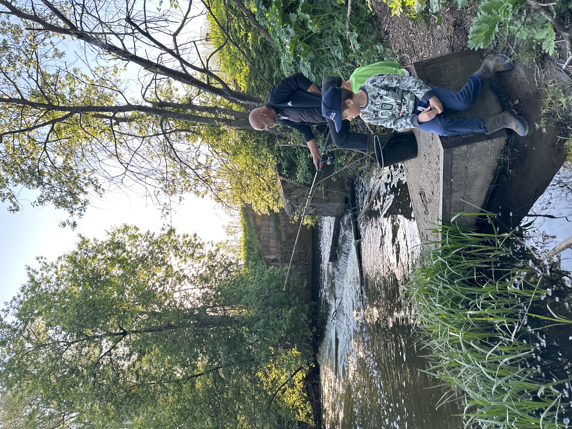 A bald man and two boys are fishing in Al'nyash, Russia. The man is holding a fishing rod, and one of the boys is sitting on a concrete structure that spans a creek or small river. The creek is flowing with some rapids near the concrete structure. The scene is surrounded by trees and vegetation.