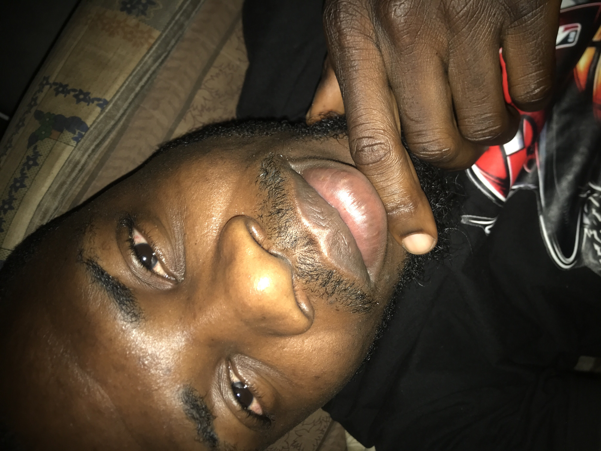 A close-up, high-angle portrait depicts a male of dark complexion, appearing to be reclining indoors and gazing directly into the camera. His face, occupying the central and upper frame, features a short, dark beard and mustache, dark eyebrows, and dark hair. His dark brown eyes exhibit a slight redness in the lower conjunctiva of his right eye (viewer's left). His full lips are glossy and slightly parted, revealing a faint smile. The skin on his forehead and cheeks reflects light prominently, indicating either an oily complexion or direct artificial illumination.
His right hand (viewer's left) is raised, with the index finger placed against his chin, just below his lower lip. The index fingernail is short and light in color. He is wearing a black garment, possibly a hoodie or t-shirt, with a partially visible graphic design in red, white, and black on the lower left portion of the image. The background consists of a patterned fabric, likely bedding, displaying beige, blue, and brown geometric or floral motifs. The lighting is artificial, creating strong highlights on the subject's skin. The setting is an indoor environment, in the city of Unknown, Unknown.