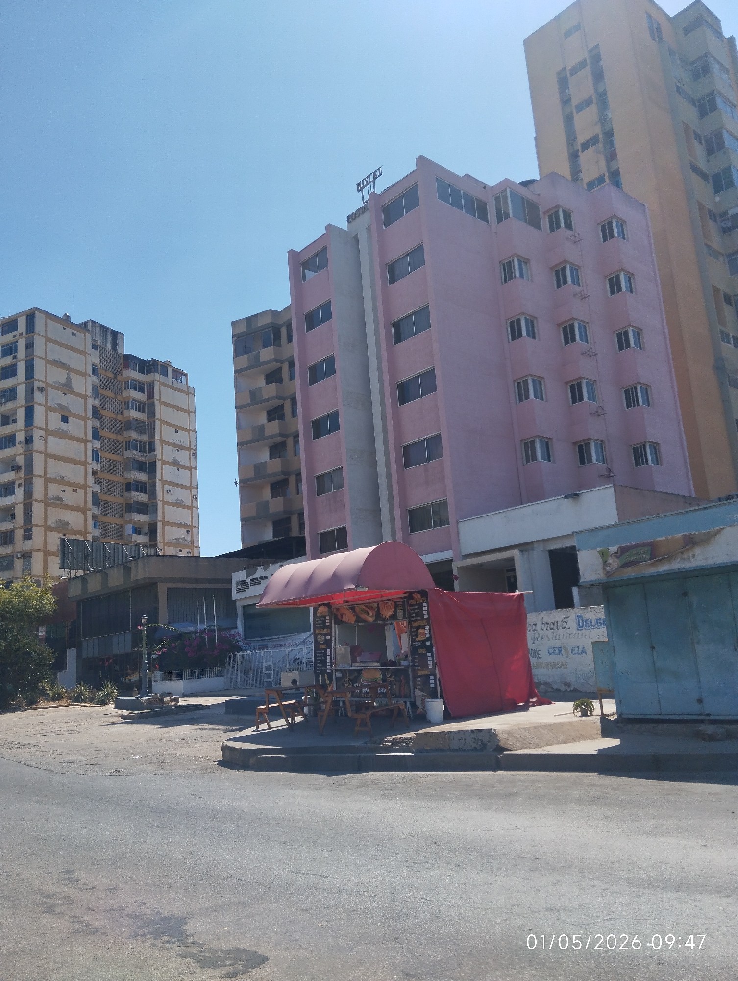 The image depicts a street scene in Porlamar, Venezuela. Several multi-story buildings, including a pink hotel, are visible. A small food stand with a red awning is set up on the sidewalk, and several chairs and tables are arranged in front of it. Graffiti is visible on a building adjacent to the food stand. The street is asphalt, and the sky is clear and blue. The date and time "01/05/2026 09:47" are stamped in the lower right corner of the image.