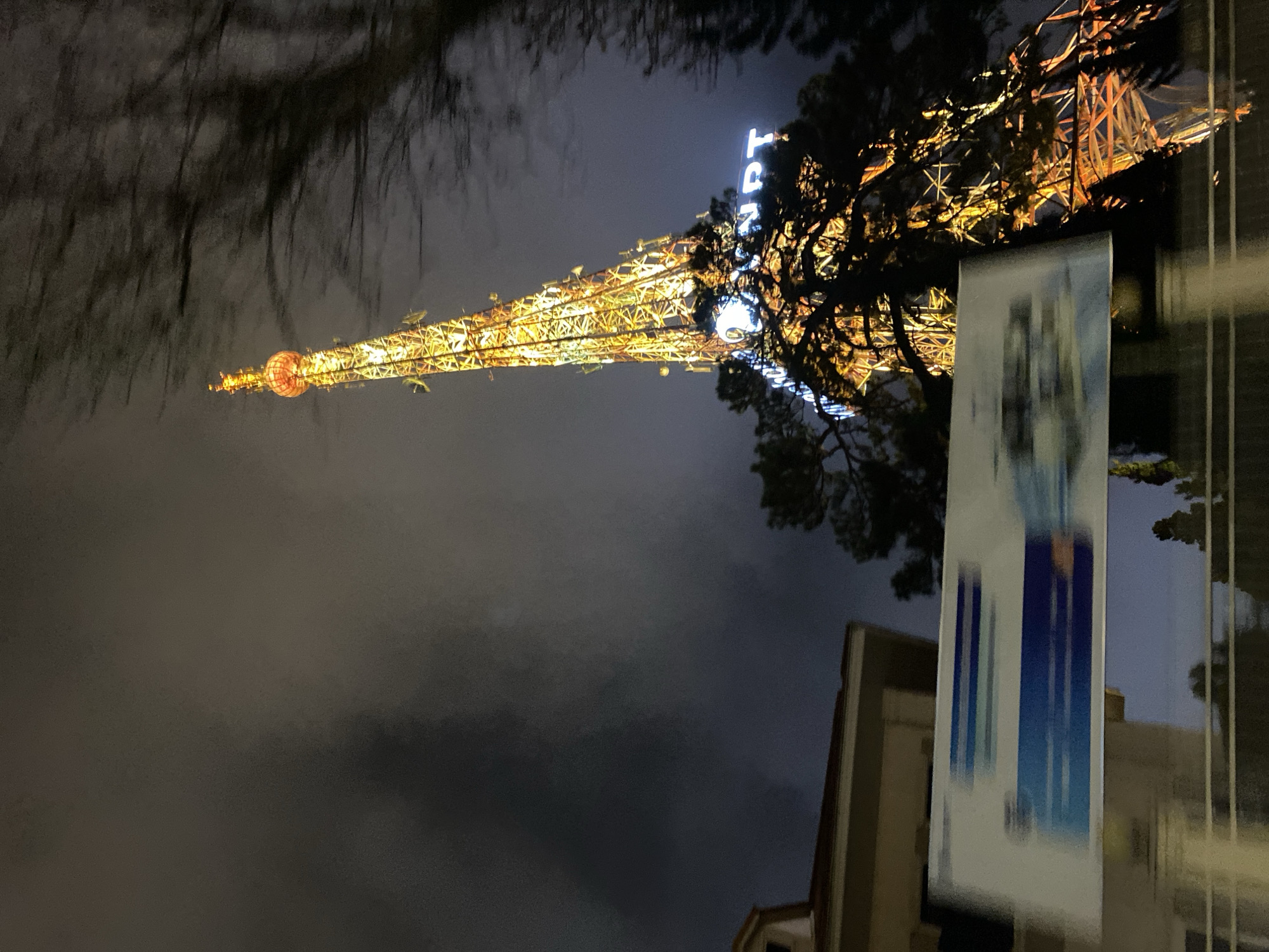 The image, originally rotated, presents a striking night scene featuring a tall, illuminated tower against a dark, cloudy sky. This structure, likely the Dalat Radio and Television Tower in Da Lat, Vietnam, stands as the central subject.
The tower's intricate lattice framework is bathed in brilliant warm yellow and orange lights, making it glow prominently in the darkness. Near its apex, a spherical or bulbous section is also brightly lit. Further down, visible against the tower's structure, are bright blue neon letters spelling out "DALAT," clearly indicating the city.
Dark, silhouetted trees with detailed branches frame the tower on both sides and intertwine with its structure, providing a natural contrast to the artificial illumination. The sky is heavily overcast, suggesting cloudy weather and contributing to the deep dark blue-gray backdrop.
In the lower right foreground, a portion of a light-colored building is partially visible. In front of this building, a vertical banner or poster, predominantly white with vertical blue stripes or gradients, stands, slightly blurring parts of the view behind it.
The scene captures the stillness of night, with no discernible activity or people present. The focus is entirely on the architectural landmark and its interplay with the natural surroundings under artificial light. The time of day is clearly nighttime, emphasized by the dark sky and the radiant glow of the tower's lights.