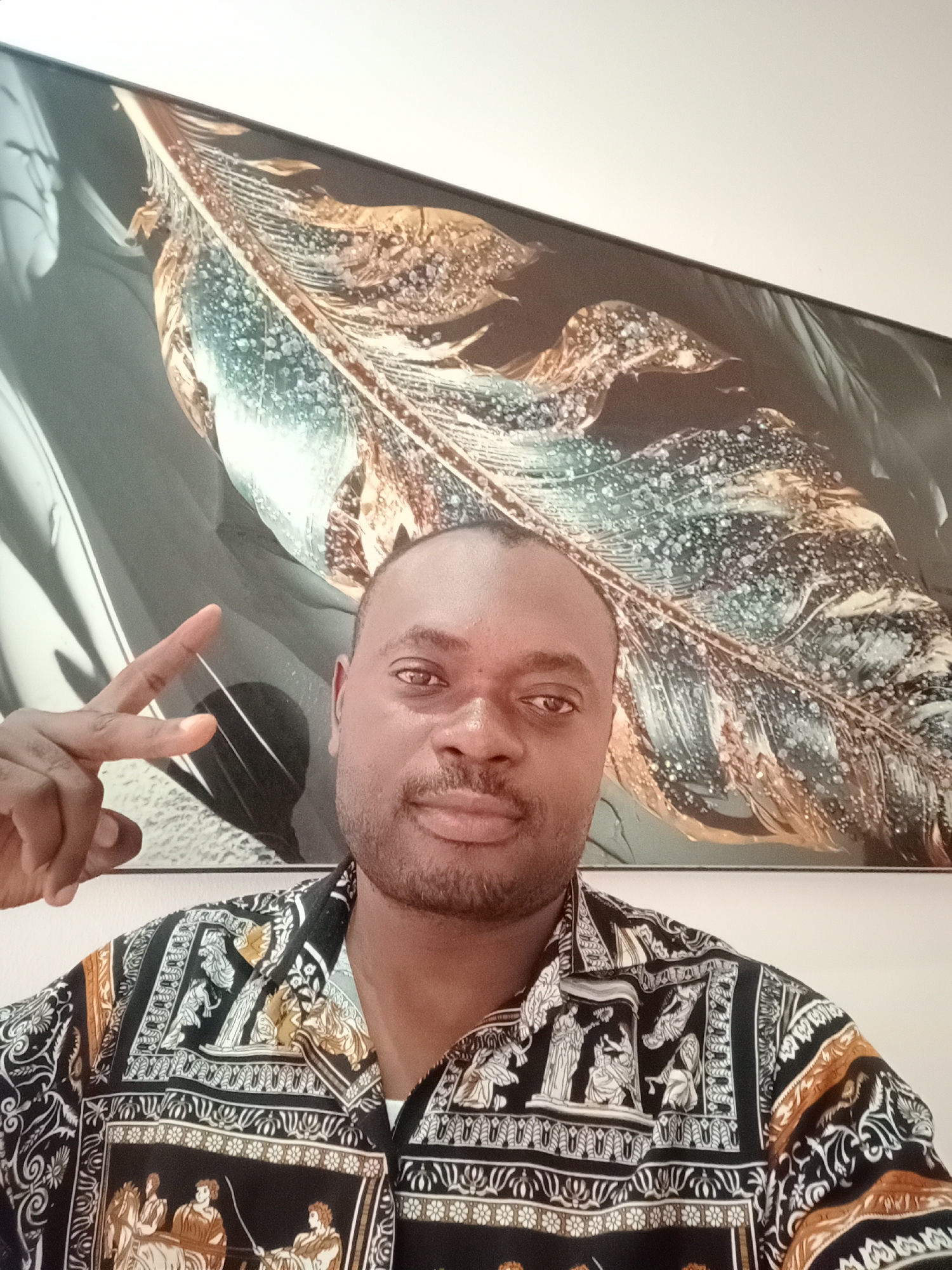 A medium shot depicts a man with dark skin and a short haircut, posing indoors against a light beige wall. He is wearing a patterned shirt with black, white, and gold colors, featuring classical Greek figures. The man is giving a "peace" or "victory" sign with his left hand. Behind him is a framed artwork of a stylized feather with a mix of teal, gold, and silver tones. The location is suggested to be in Gwarinpa, Nigeria. The photo appears to be a selfie or taken at close range.