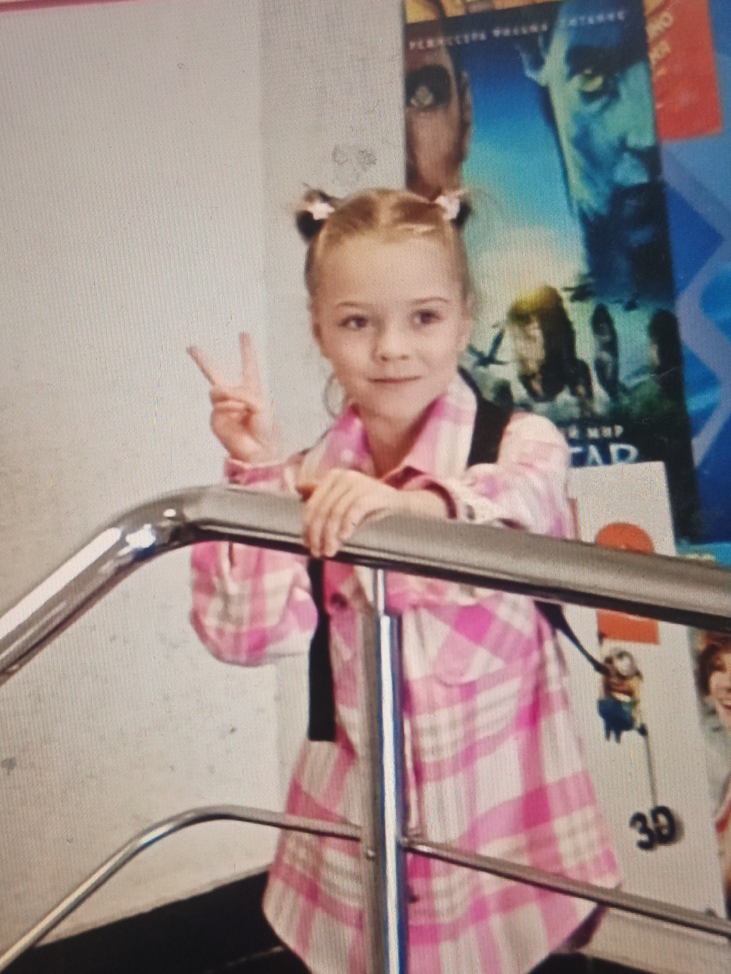 A young blonde girl with pigtails is standing and making a peace sign with her right hand. She is wearing a pink and white plaid shirt with a black backpack strap visible. She is leaning on a silver railing. Behind her are movie posters. The poster on the far right has "3D" written on it. The environment appears to be indoors, possibly a cinema lobby, with a textured wall on the left. The image quality is low, and there is significant pixelation.