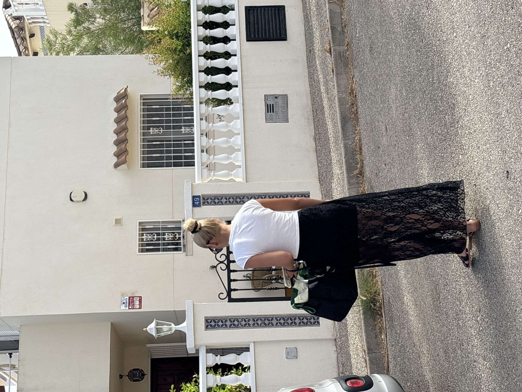 A blonde woman, viewed from behind, stands on an asphalt street in front of a cream-colored two-story house with white railings and a tile roof. She wears a white t-shirt, a black lace maxi skirt, and black sandals. In her left hand, she holds a black tote bag with green accents. The woman has her hair styled in a bun. The house has decorative tiled pillars on the left and a dark metal gate. A silver car is partially visible on the left side of the frame. The street is lined with a concrete curb and dry vegetation. A sign on the house reads "AGUA CONTADOR LLAVE DE PASO," indicating a water meter and shut-off valve. Another sign, a red and white alarm sticker, is visible near the entrance. The house number "49" is displayed on a blue plaque. The scene is illuminated by natural daylight, suggesting it is daytime.