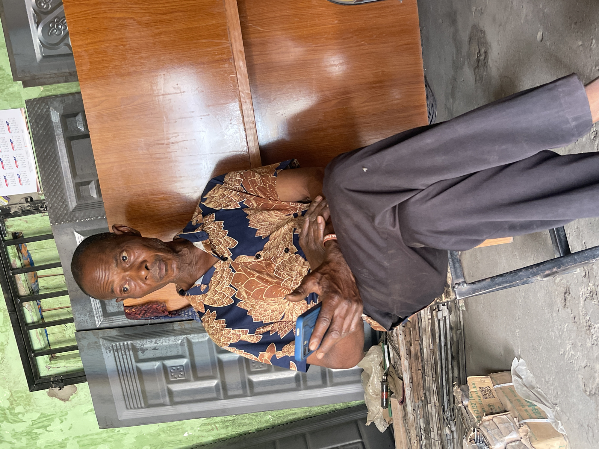 The image shows an older Black man with short, cropped hair, wearing a patterned brown and blue shirt and dark pants, sitting at a wooden table in what appears to be a workshop or warehouse setting in Zaria, Nigeria. He is holding a blue smartphone in his hands. The background features stacks of metal materials and boxes of welding materials. A calendar hangs on a wall in the upper left corner, partially obscured. The area appears to be a business that manufactures or fabricates metal doors and gates.
