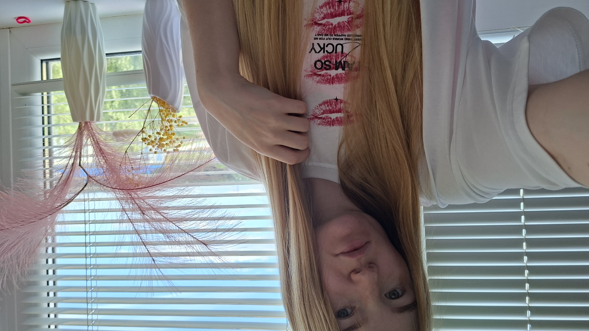 A young adult female with long, straight, reddish-blonde hair is depicted in a selfie-style photograph taken indoors in Chaikovsky, Russia. She wears a white short-sleeved t-shirt adorned with multiple red lipstick kiss marks and the prominent black text "I AM SO LUCKY," with smaller text below reading "EVERYTHING WORKS OUT FOR ME" and "MIRACLES HAPPEN TO ME DAILY." Her right hand is raised to her chest, gently touching a strand of her hair. Her fair complexion shows visible freckles on her nose and cheeks, and her eyes are blue-green with a neutral expression.
Behind her, a window fitted with horizontal white Venetian blinds allows natural daylight into the room. Through the partially open blinds, a clear blue sky with some distant green foliage is visible. On the white windowsill to the right of the woman, two decorative vases are present. The left vase is white and textured, containing a cluster of small, dried yellow flowers. The right vase is taller, with a gradient from pale mint green to white and a similar textured pattern, holding several tall, delicate, feathery pink plumes. A small, bright pink elastic band rests on the far right of the windowsill. The scene is brightly illuminated.