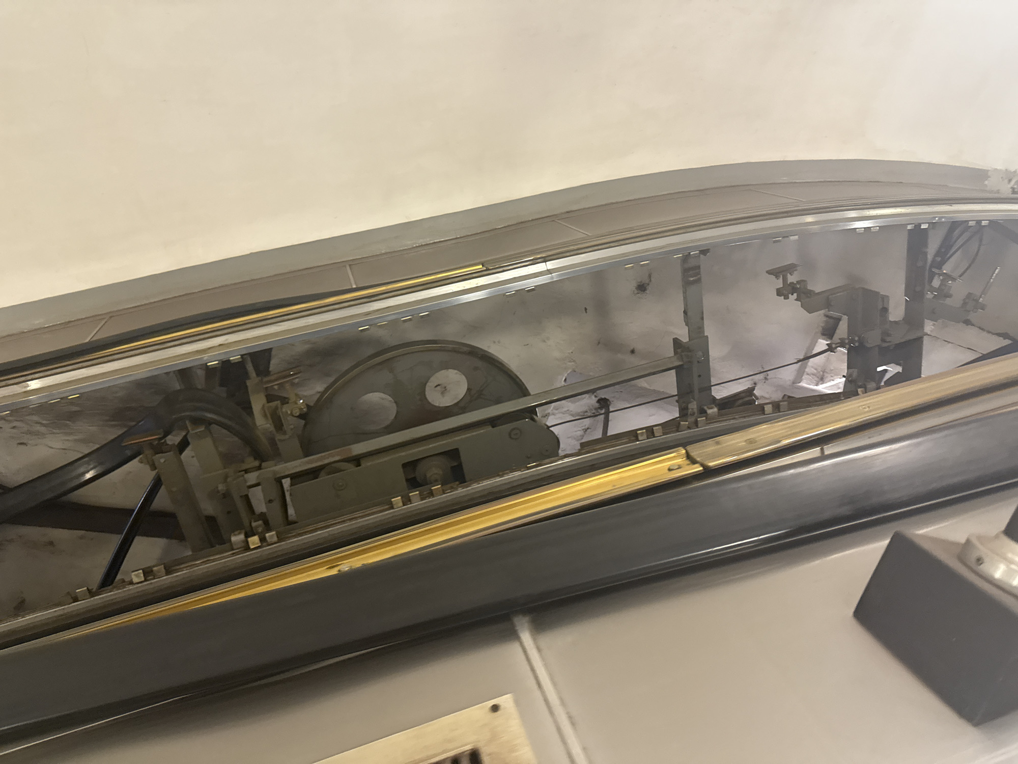 The image shows the exposed mechanical workings of an escalator. The interior is visible due to a removed or open panel. The components include a large metal wheel, various rods, and other mechanical parts. The image is taken from a low angle, looking up into the machinery. Based on the appearance, this is likely a maintenance view of an escalator system.
While the image itself doesn't explicitly provide location data, it strongly resembles the style of escalator systems in the Moscow Metro, Russia, known for their deep-level stations and elaborate escalators. Without additional location cues, this remains a highly probable but unconfirmed identification.