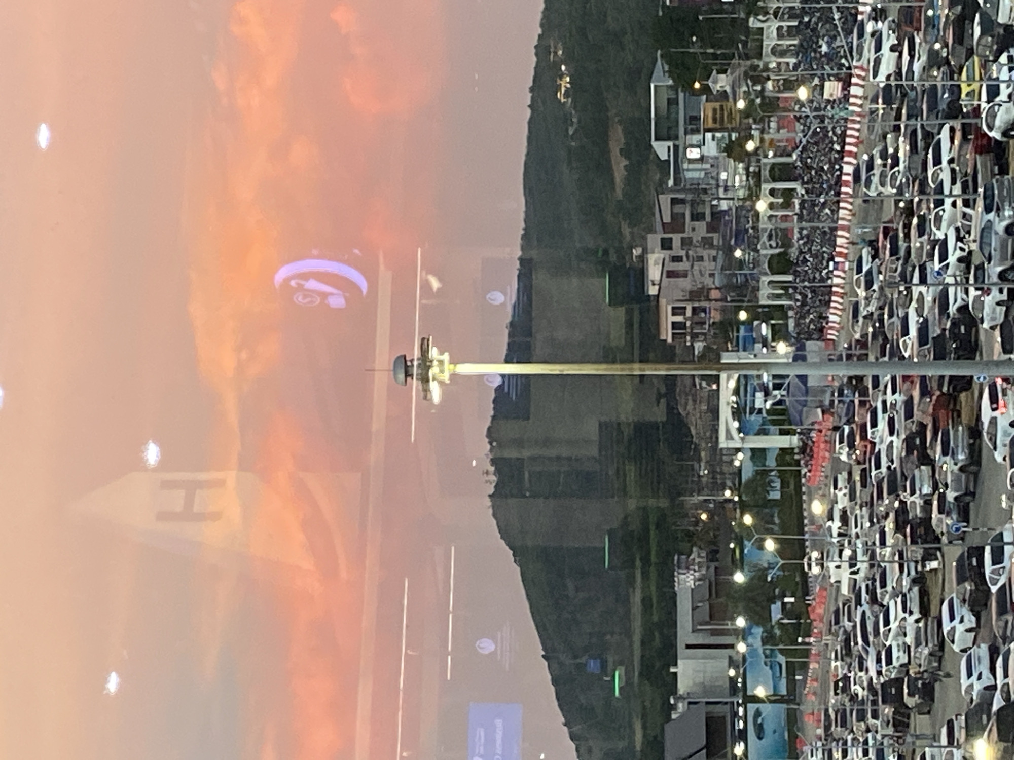 The image depicts a view from inside the S43 Hotel in Mai Khao, Thailand, overlooking a crowded outdoor event. The sky shows a vibrant sunset with orange and pink hues. A mountain range is visible in the background. In the foreground, there are buildings and a large gathering of people separated from a large parking lot with numerous parked cars. Reflected in the window are logos, including one with the letter "H" and another with a phone and sound icon.