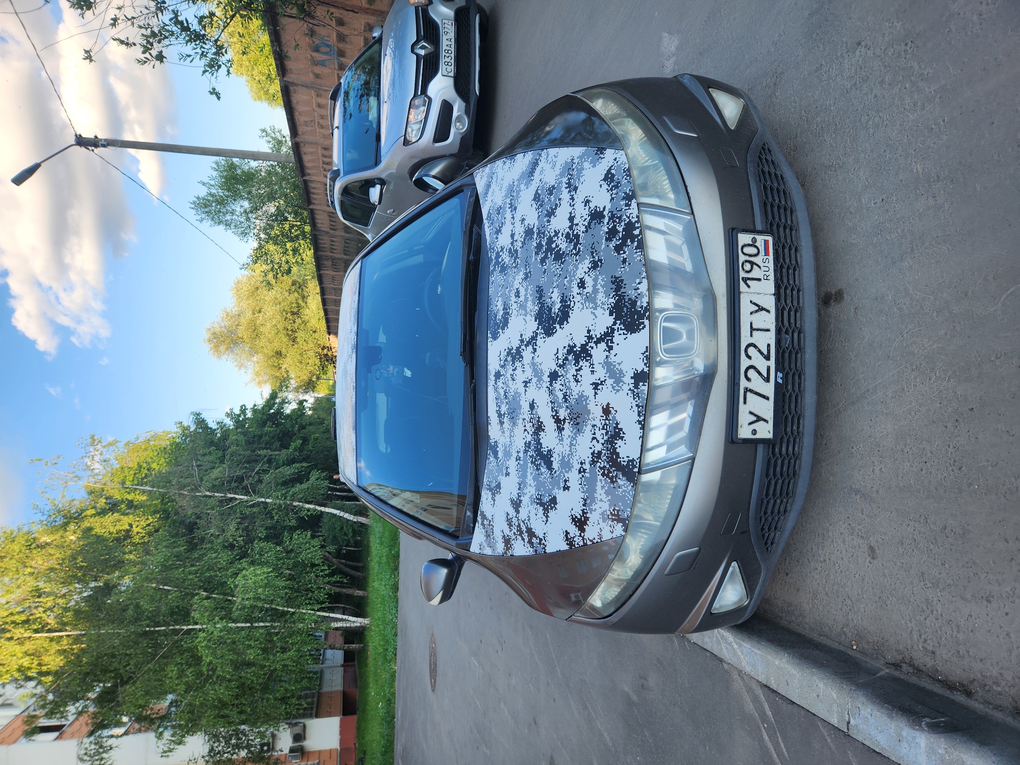 The image shows a car parked on a street in Vniissok, Russia. The car has a digital camouflage pattern on its hood and a license plate that reads "Y722TY 190 RUS". Behind this car is another car, a silver Renault Duster with the license plate "C838AA 97". The scene is set on a paved road with a concrete curb. Tall green trees and foliage are visible to the left of the cars, and a wall with graffiti is in the background. The sky is blue with white clouds, suggesting it is daytime. The lighting indicates it is likely late afternoon. There are no people visible in the image. The overall mood is calm and ordinary, depicting a typical street scene.