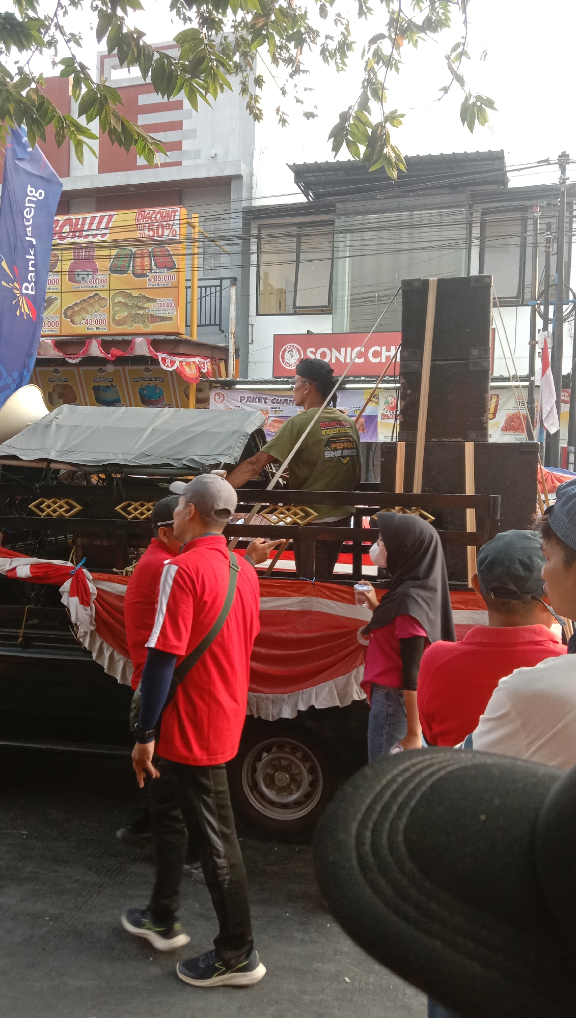 The image captures a street scene in Jatisawit, Indonesia, likely during a daytime event given the bright sunlight and clear sky. In the foreground, several individuals are visible from behind, suggesting they are spectators or participants in a procession or gathering. One person in a red shirt and black pants is facing away from the camera, appearing to interact with someone to their right. Another person in a red shirt is partially visible to the left.
A key element of the scene is a decorated truck, serving as a mobile stage or float. It is adorned with red and white fabric draped around its sides and features a large speaker system mounted on top. A man in a green shirt is standing on the truck, possibly operating equipment or addressing the crowd. Various banners and signs are displayed on buildings in the background, indicating commercial establishments. One sign clearly shows "SONIC CH" which could be part of a business name. Another sign depicts food items with prices, suggesting a food-related promotion or sale. The presence of overhead wires and the architectural style of the buildings point to an urban or semi-urban environment in Indonesia. The overall impression is of a lively, community-focused event taking place outdoors.