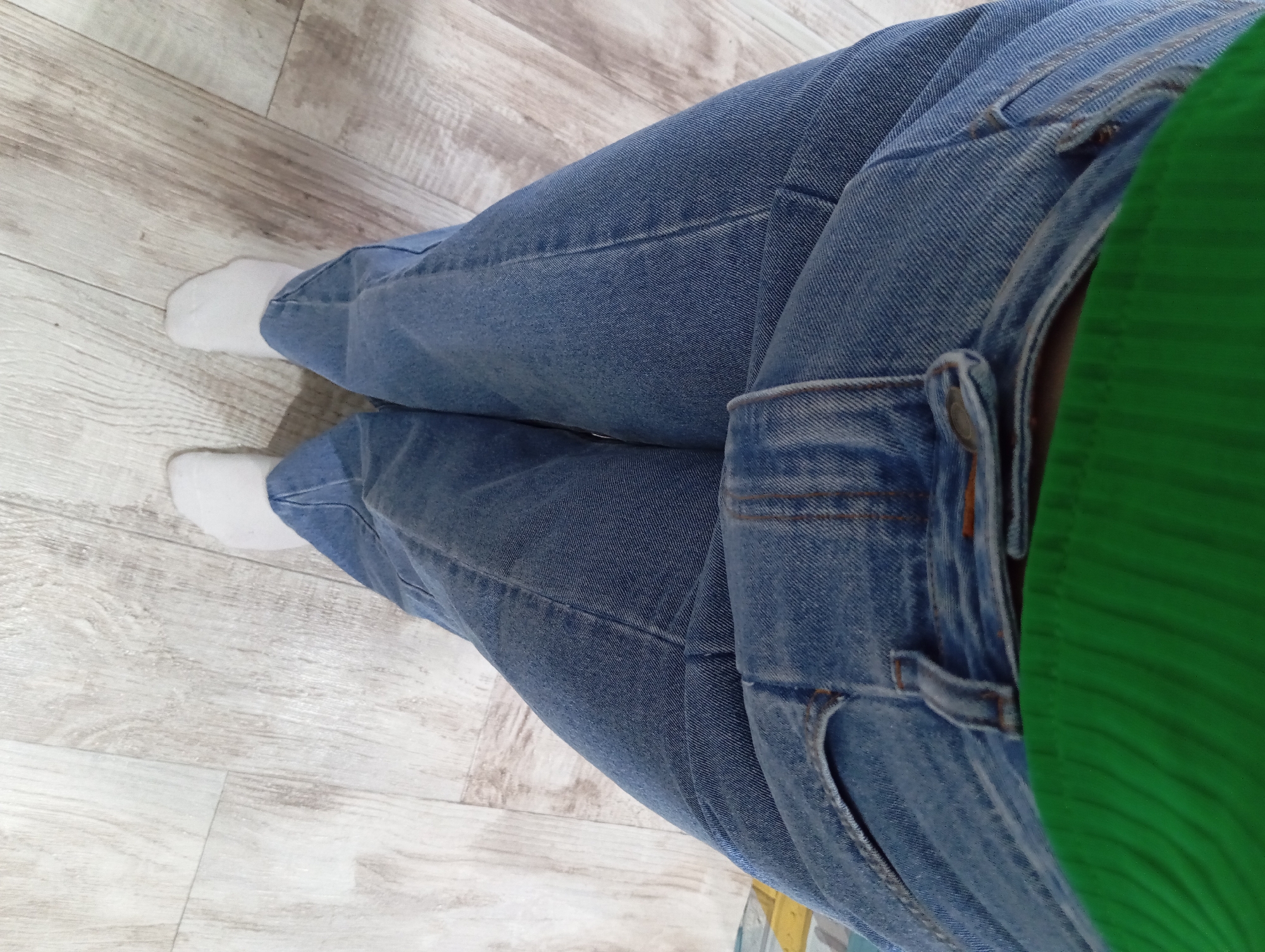 The image depicts a person's legs wearing blue jeans and white socks. The person is standing on a light-colored wooden floor. The top of a green garment is visible at the right edge. The location is possibly inside a building, but the context is limited. There is no information to indicate this is in Yurgamysh, Russia.
