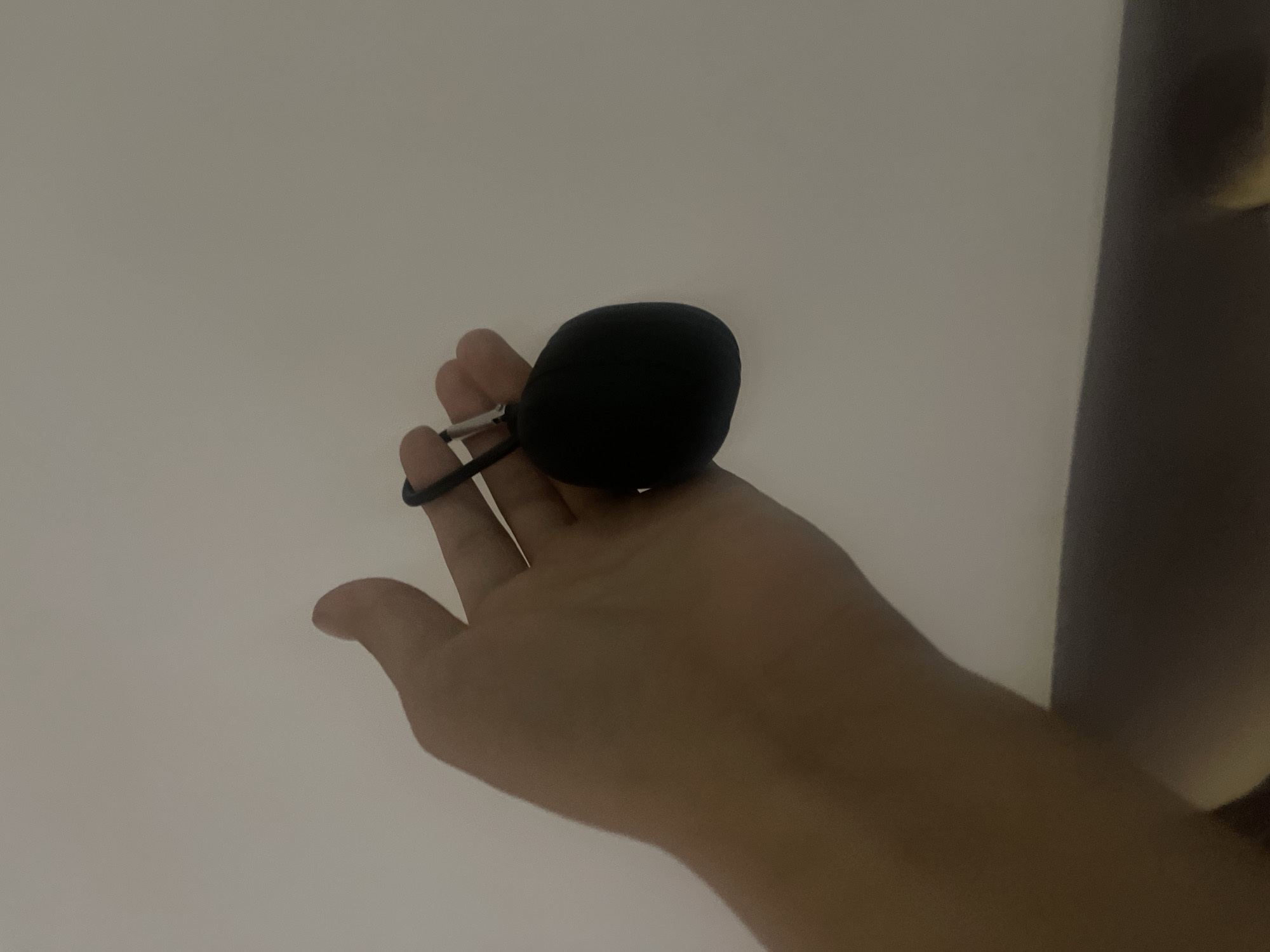 The image shows a person's hand holding a small, black, round object. The object appears to be a microphone windscreen or a small, portable speaker. A thin black cord is attached to the object, with a small metal clasp or ring. The hand is positioned palm-up, and the fingers are slightly curled around the object. The background is a plain, light-colored surface, possibly a wall or ceiling, with a soft, diffused light. The lighting suggests an indoor setting. There is a hint of a darker, vertical element on the right side of the frame, which could be furniture or a doorway. No text is visible in the image. The focus is on the hand and the object it holds. The overall impression is one of a simple, everyday moment.