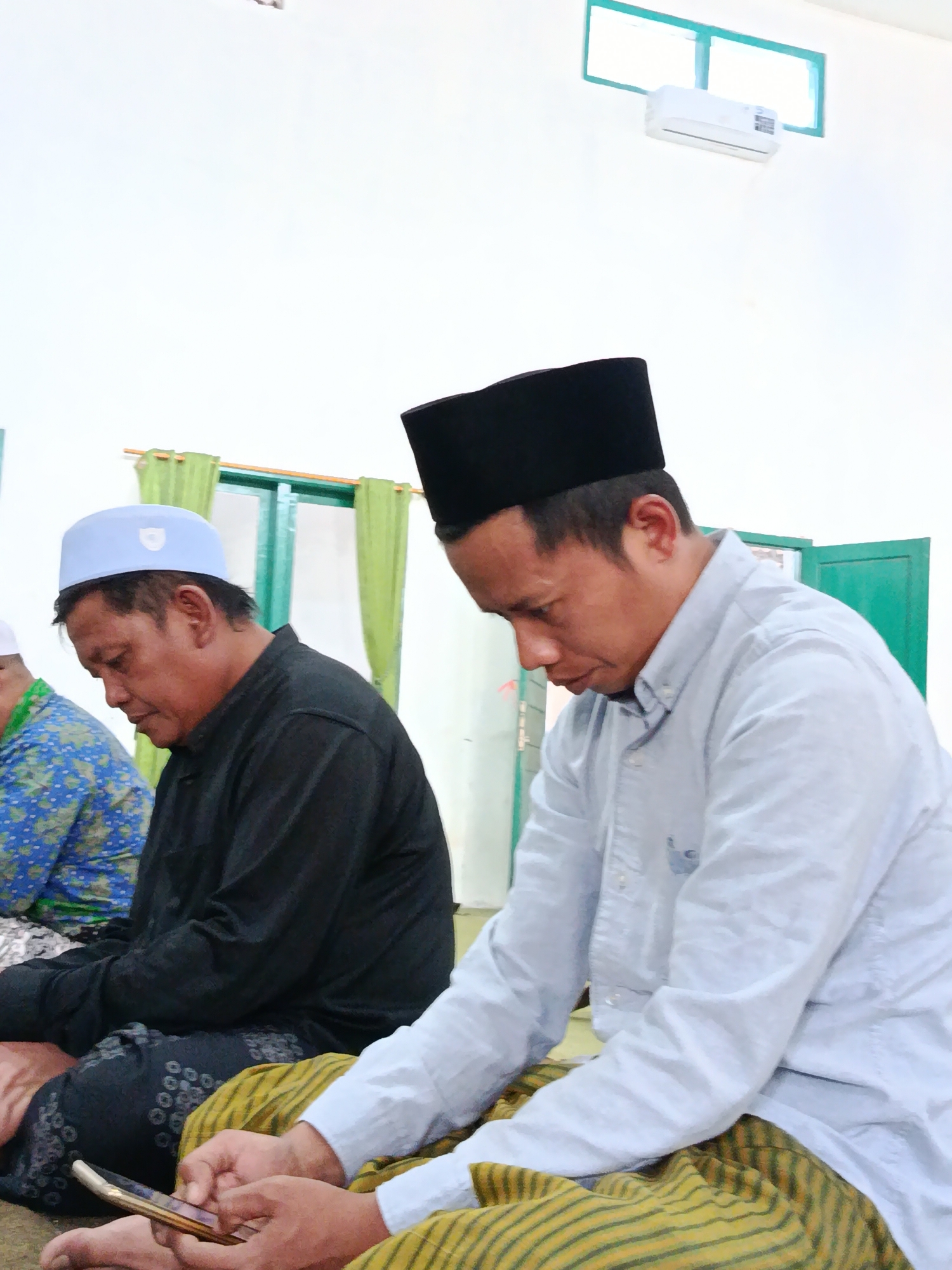 Two men are seated on the floor in a room. The man on the left is wearing a black long-sleeved shirt and a light blue cap. The man on the right is wearing a light blue long-sleeved shirt and a black cap. Both men are looking down. The man on the right is holding a smartphone. In the background, there are green curtains and a window with an air conditioner. This scene likely takes place in a mosque or a prayer room in Bondowoso, Indonesia, given the attire of the men and the presence of prayer rugs.