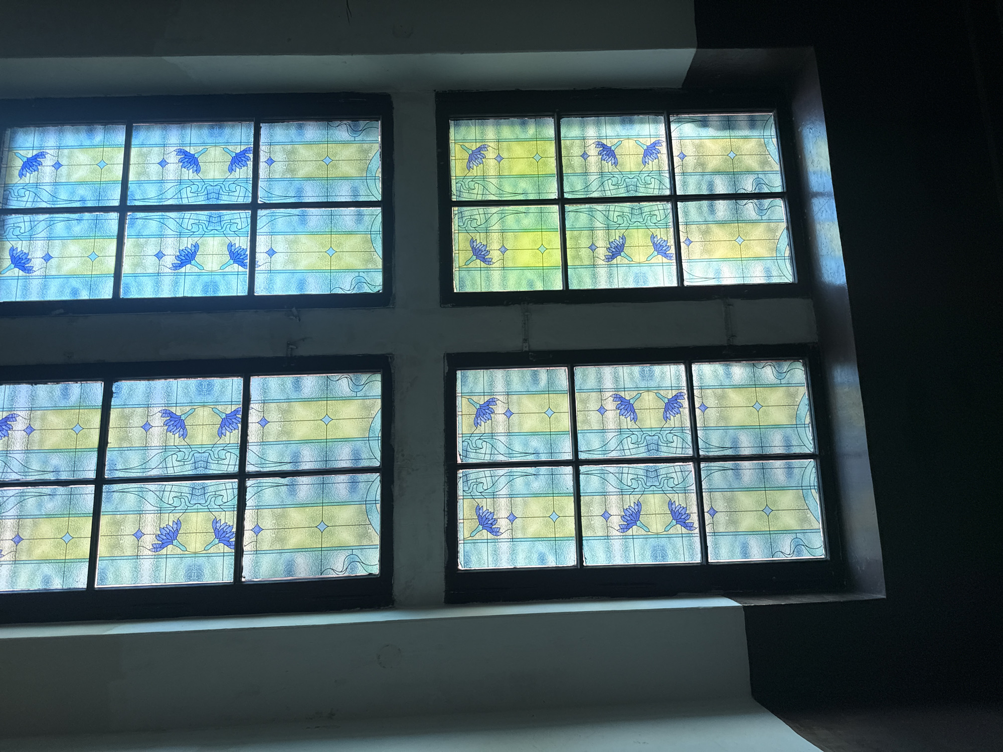 The image depicts four stained glass windows arranged in a square formation. Each window is divided into six panes by dark frames. The stained glass features a repeating pattern of blue floral designs and wavy lines against a background of blue and yellow hues. The windows appear to be located indoors and are positioned above a light-colored, stepped ledge. The setting is the city of Karpaty, Ukraine.