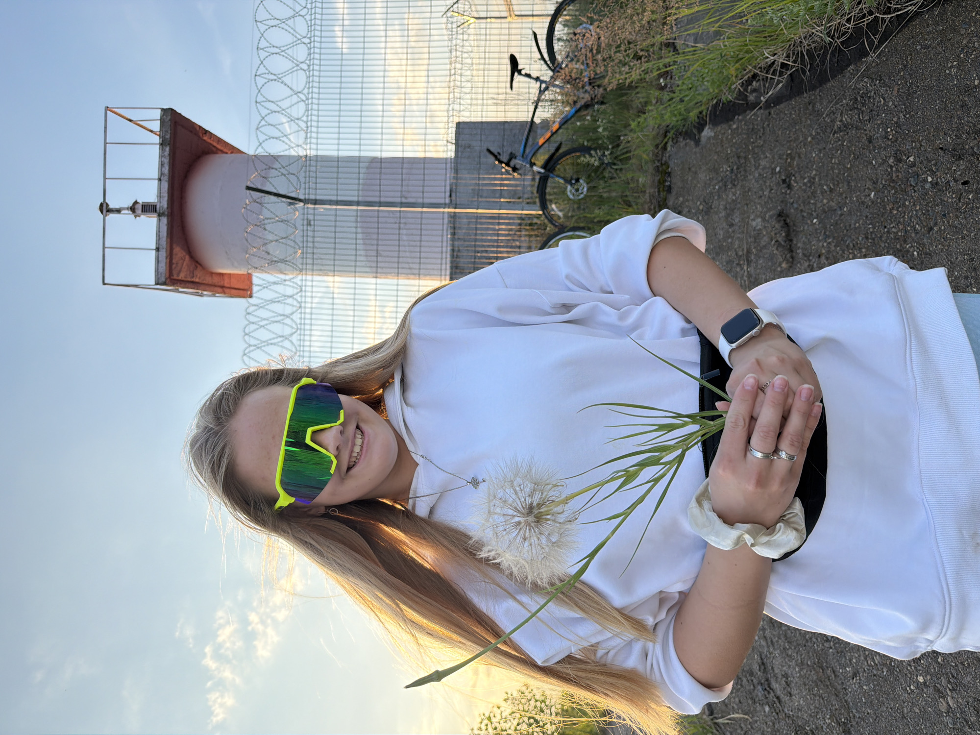 A young blonde woman with long hair wears bright neon green sunglasses and a white hoodie. She is holding a large dandelion. She has a black fanny pack around her waist and wears rings on her fingers and a smartwatch on her left wrist. In the background, a lighthouse and a fence are visible, along with two bicycles leaning against the fence. The scene is outdoors, with grass and a concrete path. The sun is setting, casting a warm glow. This location is consistent with the area near the Kama River dam in Chaikovsky, Russia, a common spot for recreational activities and picturesque views.