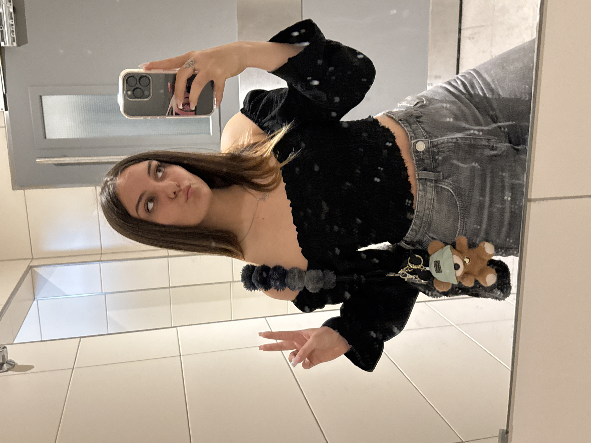 A young woman is captured taking a mirror selfie indoors, likely in a public restroom or changing room in Sofia, Bulgaria. The image is rotated 90 degrees clockwise, showing her full-length reflection. She has long, dark brown hair and is looking upwards with a slight pout on her face.
She is dressed in a black, off-the-shoulder top with puffy sleeves and a textured fabric that appears to have small white speckles. Her outfit is completed with distressed grey jeans. A series of dark grey/black fuzzy pompoms are visible as an embellishment on her top or an accessory around her waist.
With her right hand, she holds an iPhone featuring a triple camera system, used to take the selfie. A ring is visible on her right ring finger, and her fingernails are painted a light, neutral color. Her left hand is making a peace sign gesture. Clipped to her jeans is a small, light brown teddy bear keychain wearing a light blue face mask. A delicate necklace with a small pendant is also visible around her neck.
The background consists of light-colored, possibly white, tiled walls, with grout lines creating a grid pattern. A frosted glass panel or window is visible in the upper left section of the reflected background. The setting suggests an indoor public space, and the overall mood is casual and posed. No visible text is present in the image.