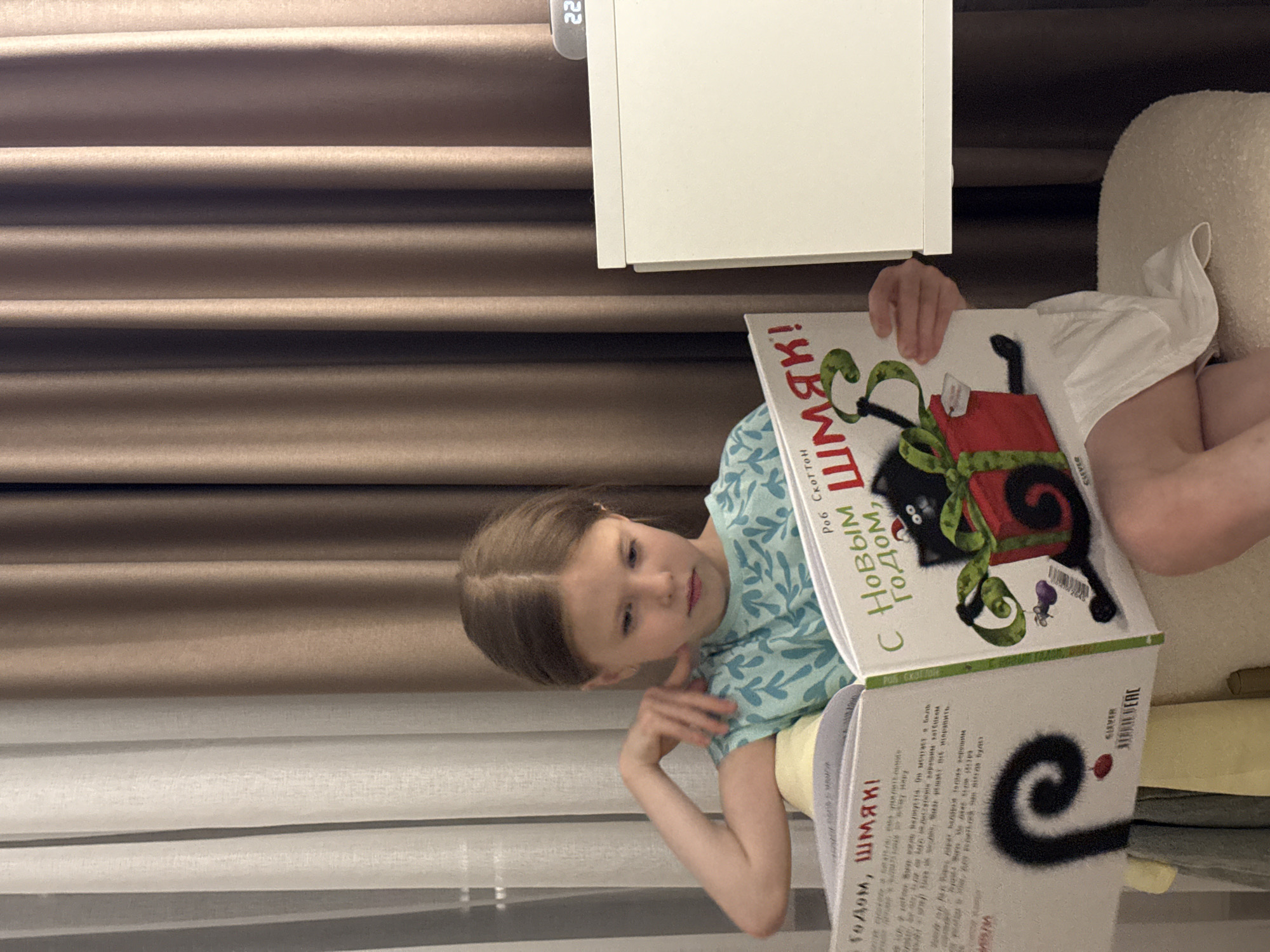 A young girl is sitting and reading a book. The book is titled "Happy New Year, Splat!" by Rob Scotton, with the title in Russian ("С Новым ШМЯК! Годом"). The girl is wearing a light blue shirt with leaf-like patterns. The environment includes curtains, a white nightstand, and a beige chair. The time on the digital display nearby is 22. The location in the city of Izhevsk, Russia, is not determinable from the image.