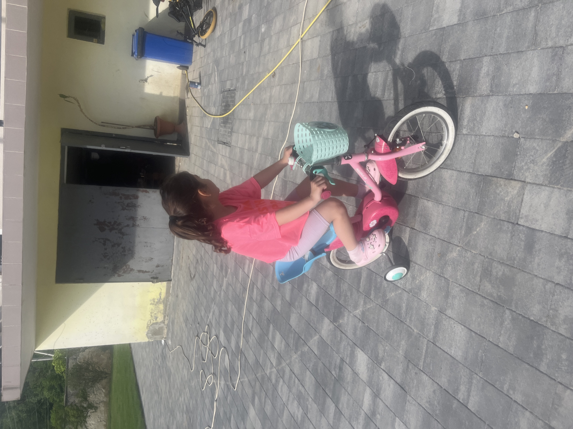 In Onokivtsi, Ukraine, a young girl with dark hair in a ponytail is riding a pink children's bicycle with training wheels. She is wearing a bright pink t-shirt and light purple shorts. The bicycle has a light blue seat and a light blue basket on the handlebars. The girl is looking to her right as she rides on a paved area made of grey interlocking stones. Behind her is a light yellow building with a large, dark, weathered garage door and an open doorway. A potted plant is visible to the left of the open doorway. Further to the right, a blue trash bin stands near a yellow hose. The ground is dry and appears to be a driveway or patio area.